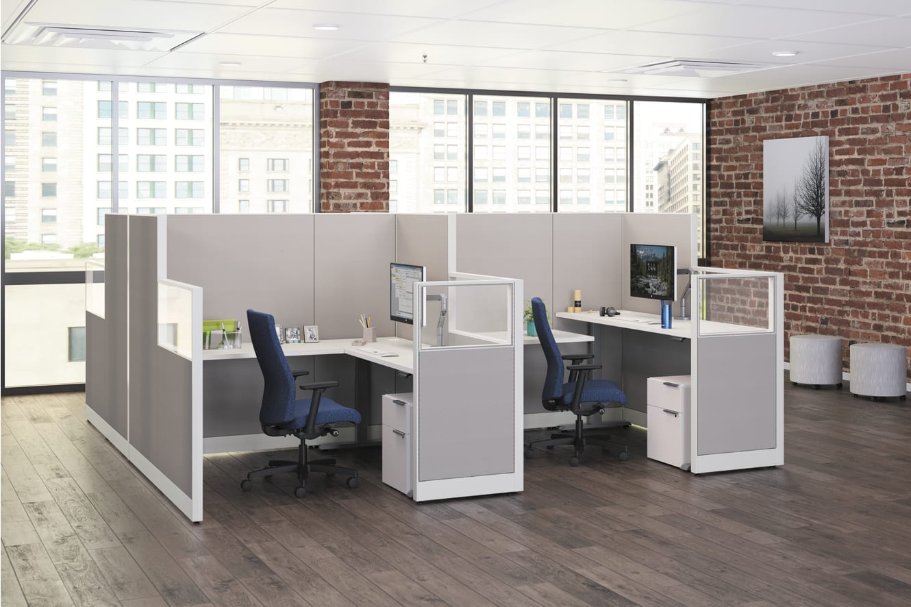 The image depicts a modern office space with cubicles, desks, and computers, set against a backdrop of large windows and exposed brick walls.