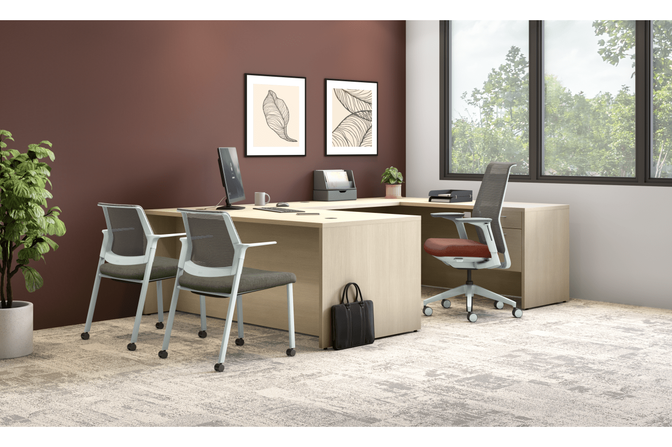 A modern and minimalist home office setup with a wooden desk, white chairs, and framed artwork on the burgundy-colored walls, set against a backdrop of large windows overlooking a lush, green outdoor environment.
