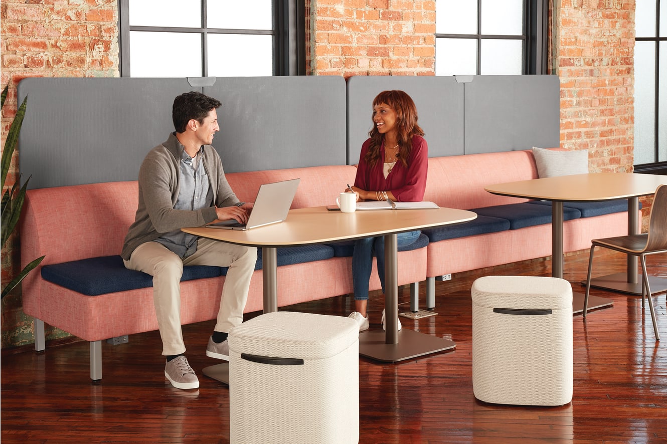 Two people, a man and a woman, are seated at a table in a modern office-like setting with brick walls and large windows in the background.