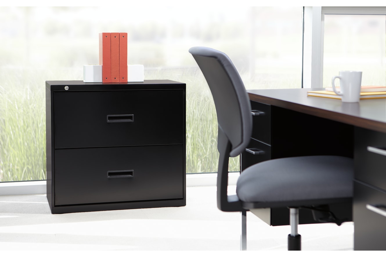 A black filing cabinet stands in the foreground, with a red folder on top, next to a swivel chair in front of a large window overlooking a grassy outdoor scene.
