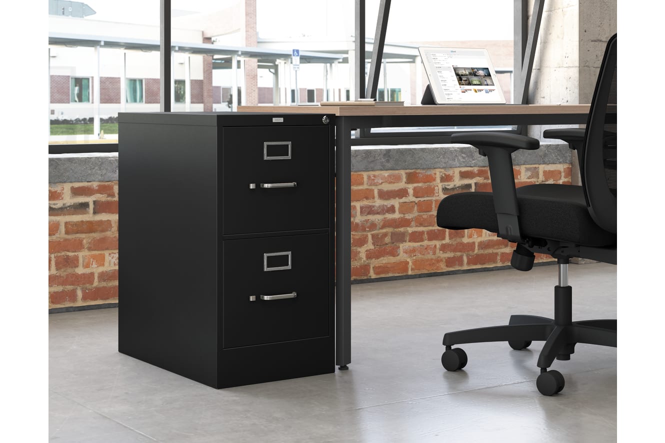 A black filing cabinet stands in the foreground, with a desk and office chair visible in the background, set against a brick wall.