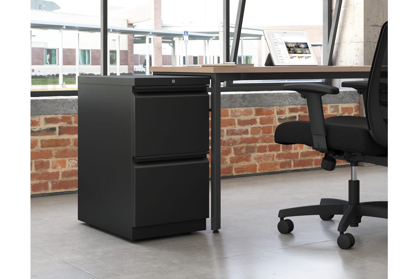 A modern office workspace with a black filing cabinet and a swivel chair in the foreground, set against a backdrop of brick walls and large windows.