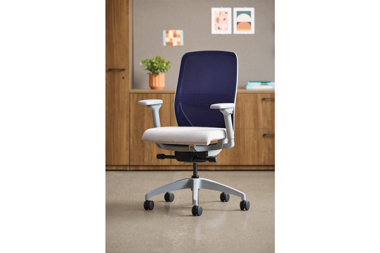 A modern, adjustable office chair with a navy blue backrest and a light-colored seat sits in a cozy, well-furnished room with wooden cabinets and framed artwork on the walls.