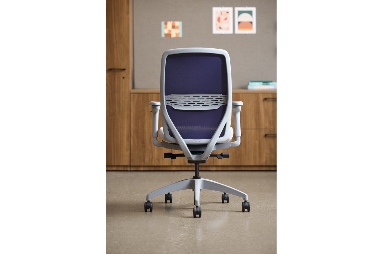A modern, ergonomic office chair with a purple mesh backrest and seat, situated in a minimalist workspace with wooden cabinets and framed artwork on the walls.