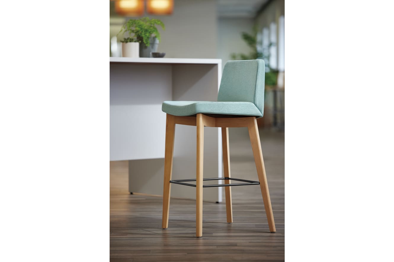 A modern, minimalist chair with a light green upholstered seat and wooden legs sits in a room with a white desk and potted plant in the background.