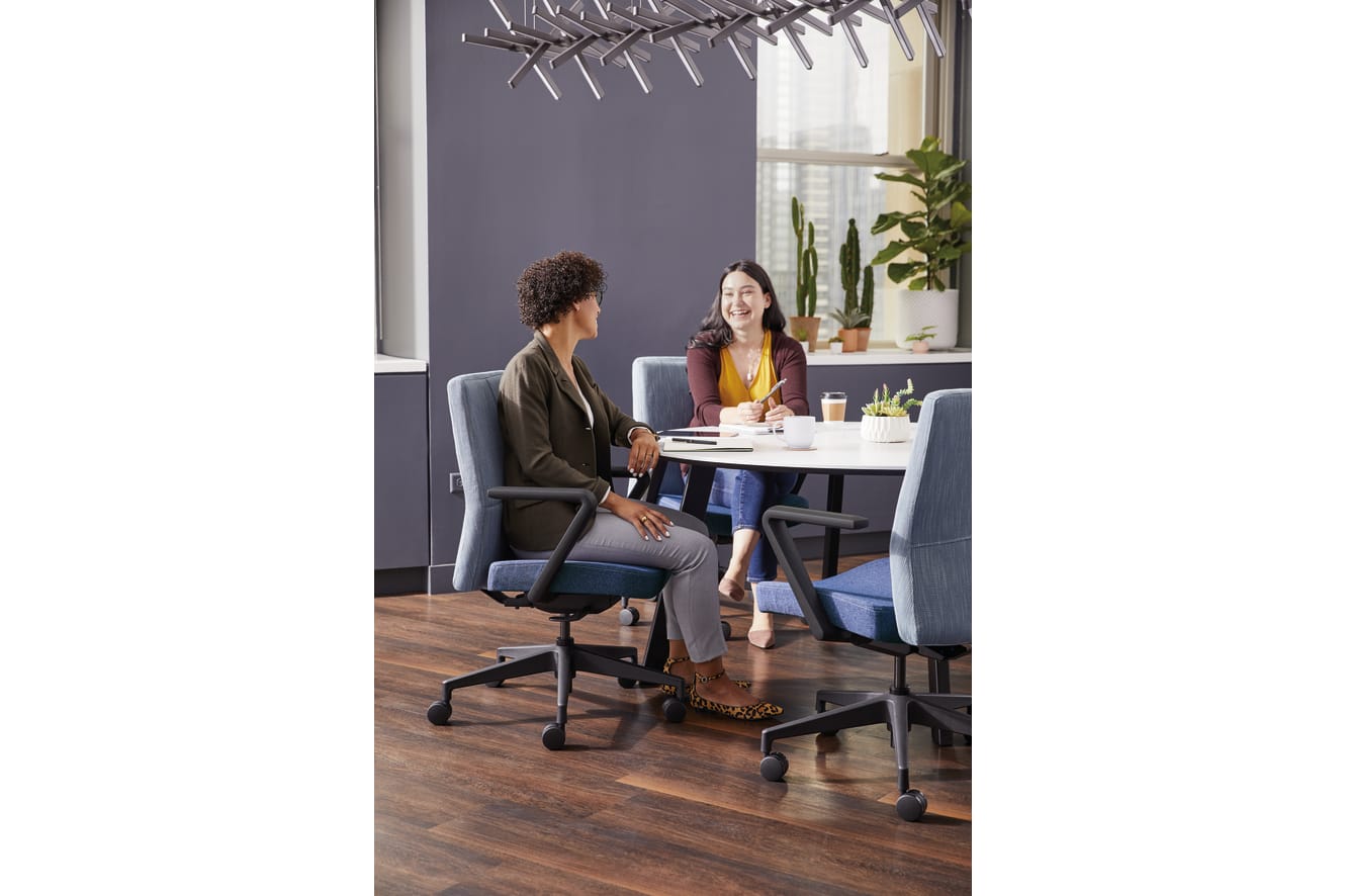 Two people, a woman and a man, are sitting at a table in an office-like setting, engaged in conversation. The background features a gray wall, potted plants, and a modern lighting fixture.
