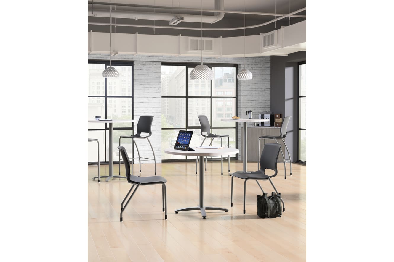 A modern, minimalist office space with desks, chairs, and a laptop on one of the tables, set against a backdrop of large windows and exposed brick walls.