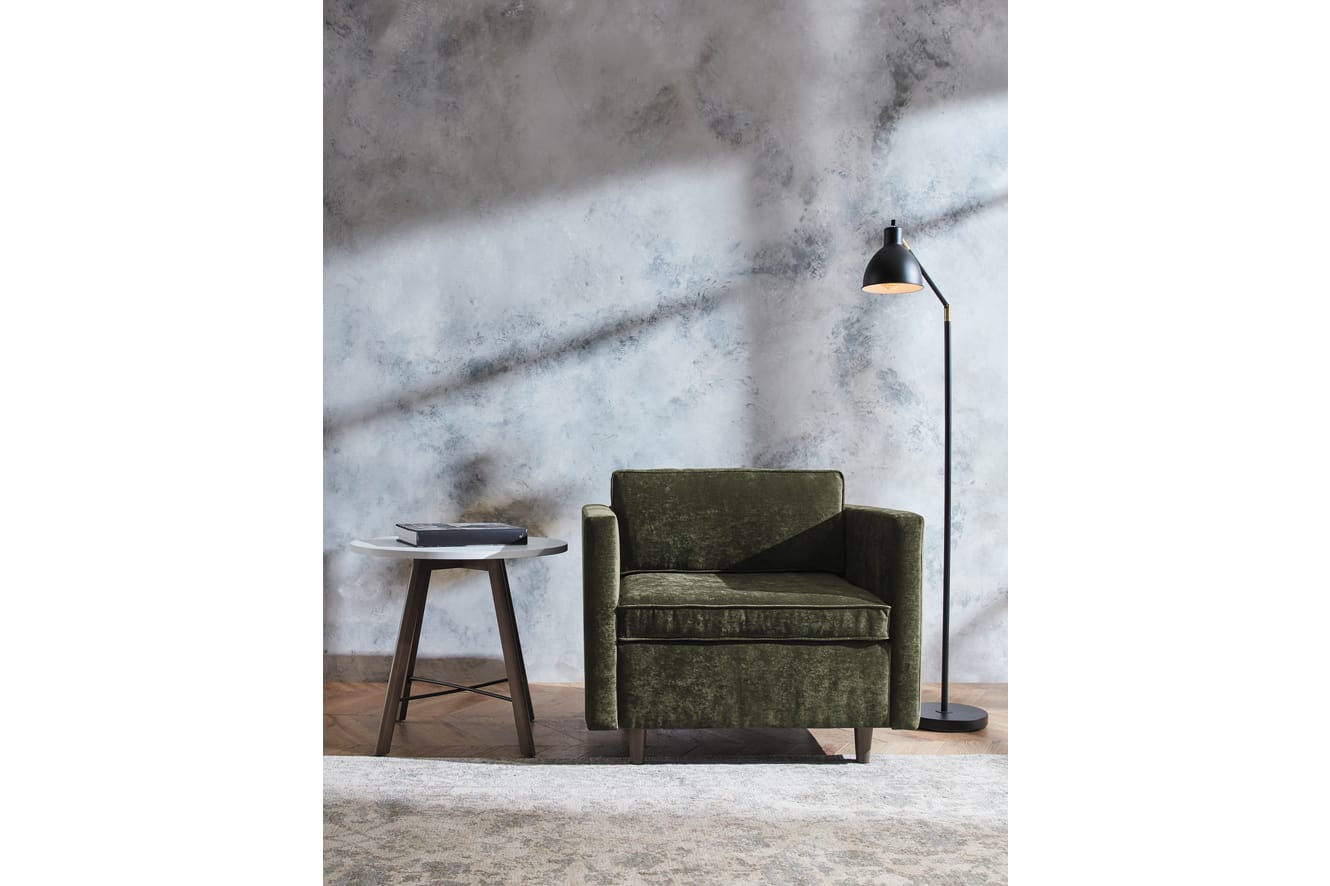 A cozy green velvet sofa sits in a dimly lit room, with a small wooden side table and a floor lamp providing a warm glow against a textured, gray wall backdrop.