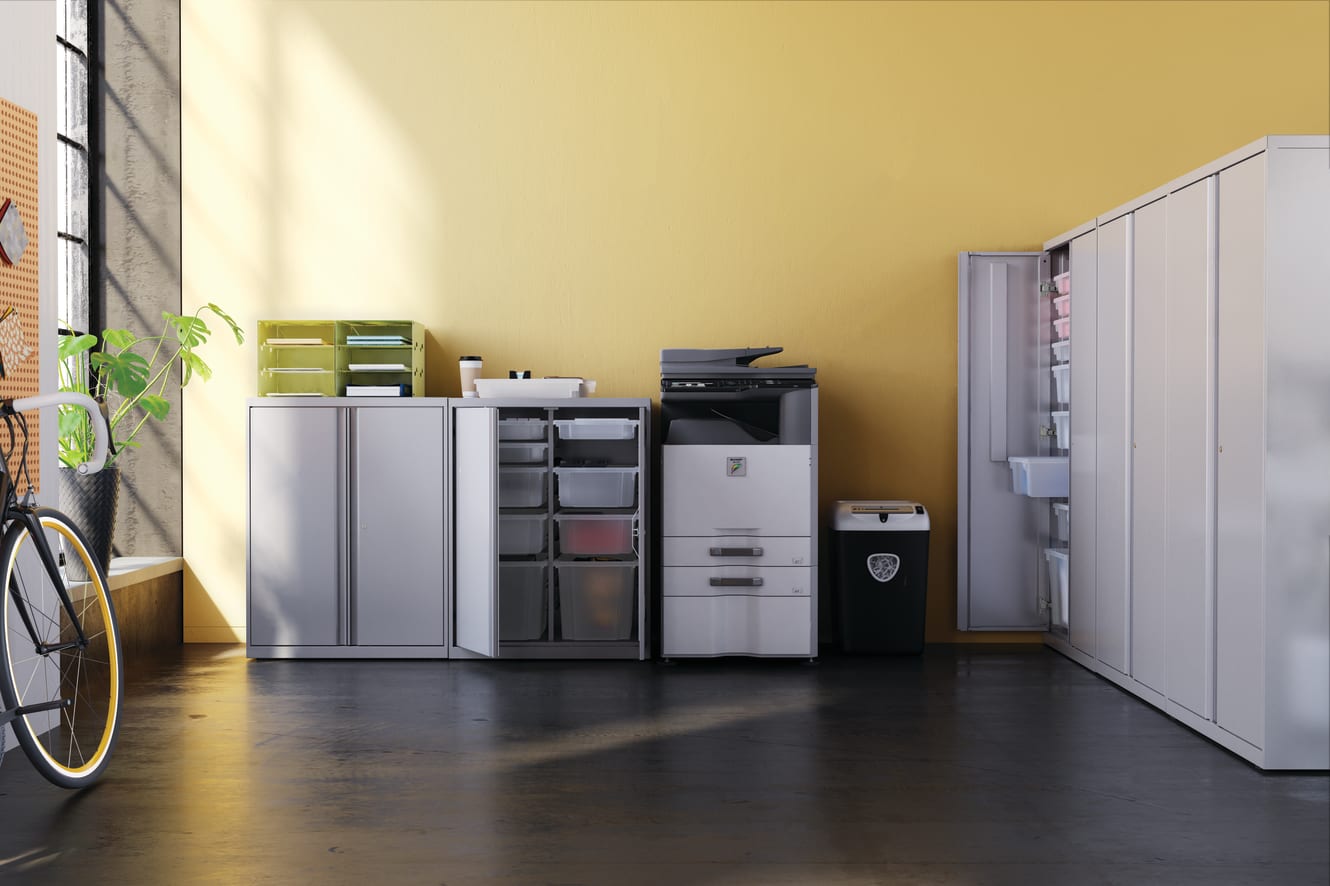 A cluttered office space with various storage units, a computer, and a bicycle visible in the foreground, set against a yellow wall in the background.