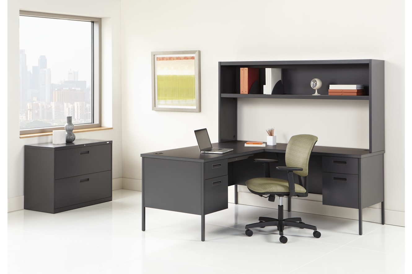A modern and minimalist home office setup with a black desk, shelving unit, and a green office chair against a white wall with a large window.