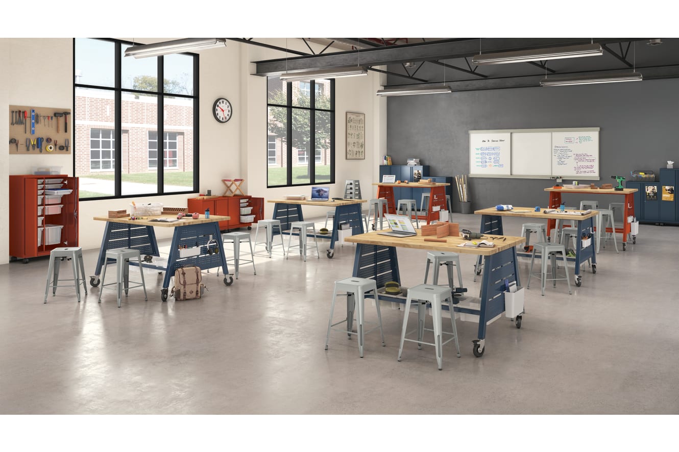A modern, open-concept workspace with various workstations, desks, and chairs arranged throughout the room. The space features large windows, exposed ceiling beams, and a whiteboard on the wall, creating a bright and collaborative environment.