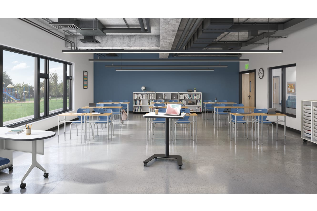 Modern classroom with flexible desks and chairs, large windows, blue accent wall, ceiling lighting, bookshelves, and teacher workstation with laptop and coffee cup.