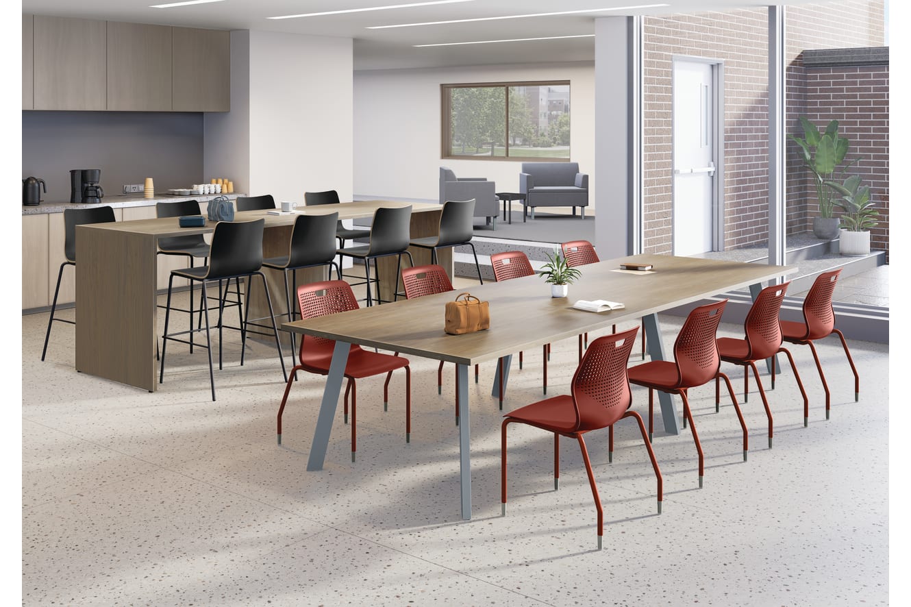 Modern collaborative workspace with long wooden tables, red and black chairs, coffee station, adjacent lounge seating, large windows, and bright contemporary lighting.