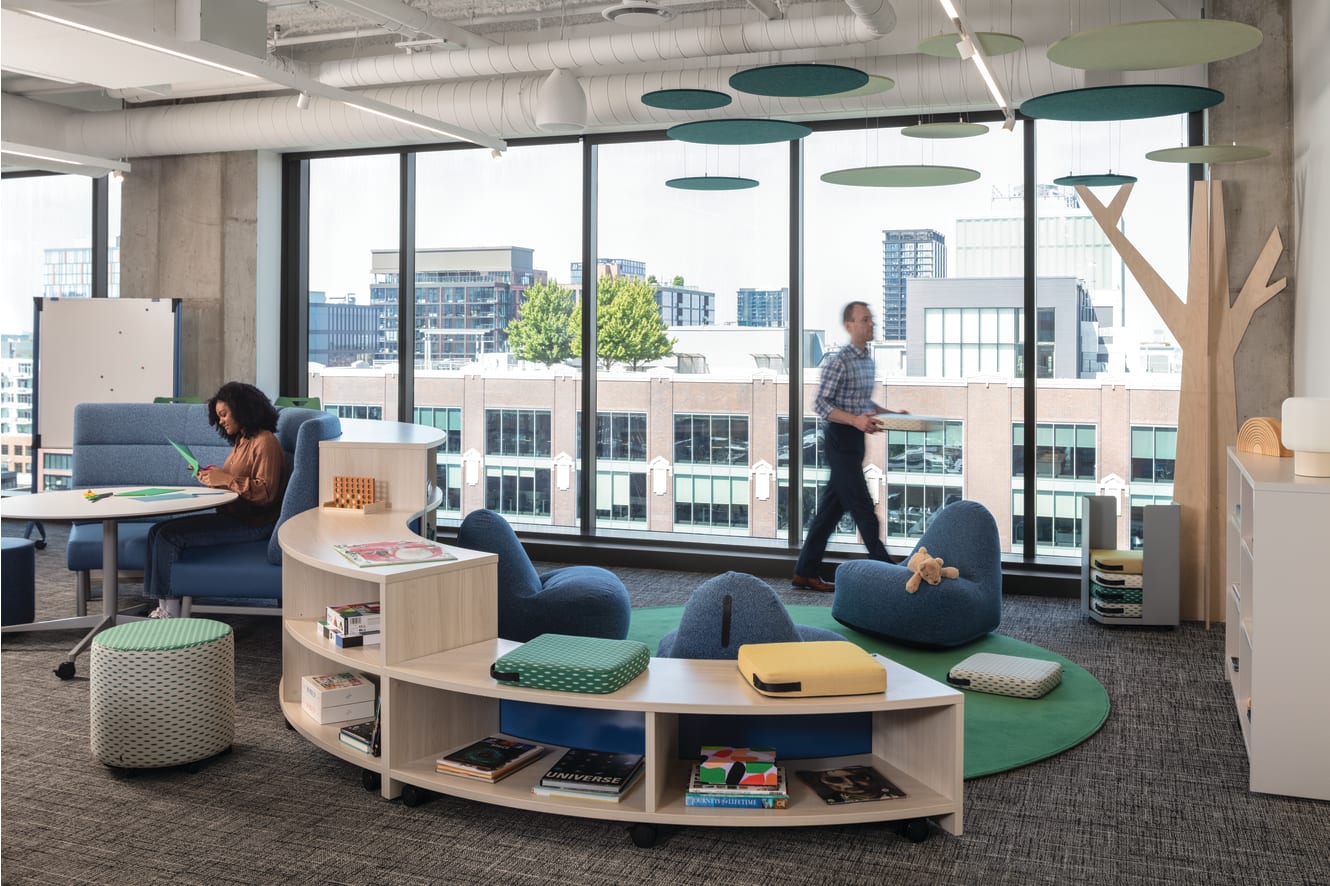 Copilot said: Flexible lounge area with soft seating, low tables, shelving, and large windows overlooking city buildings and trees.
