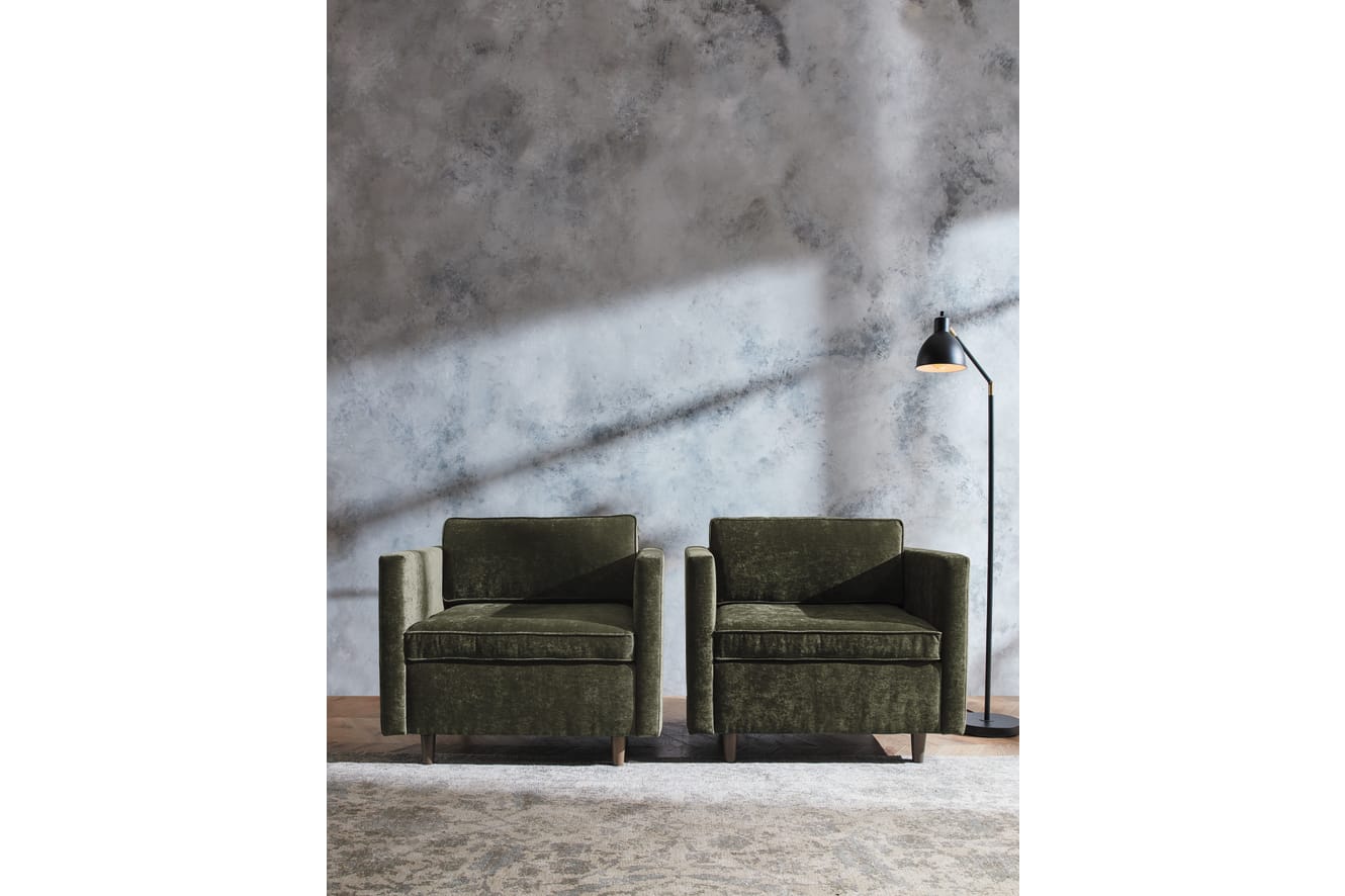 Two olive green leather armchairs sit in a minimalist room with a textured concrete wall in the background and a tall floor lamp illuminating the space.