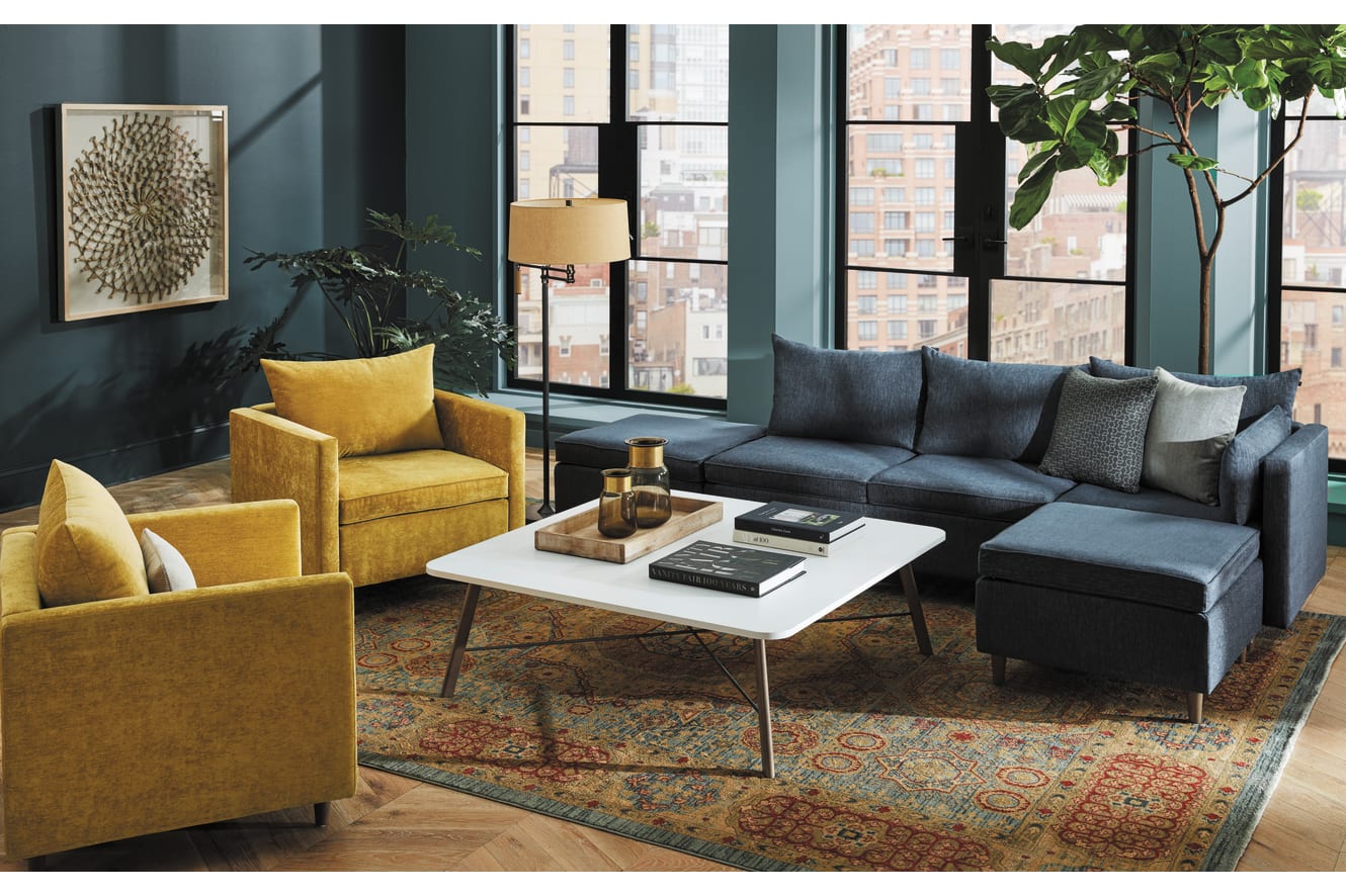 A cozy and stylish living room with a gray sectional sofa, yellow armchairs, a white coffee table, and a patterned area rug, set against a backdrop of large windows and lush greenery.