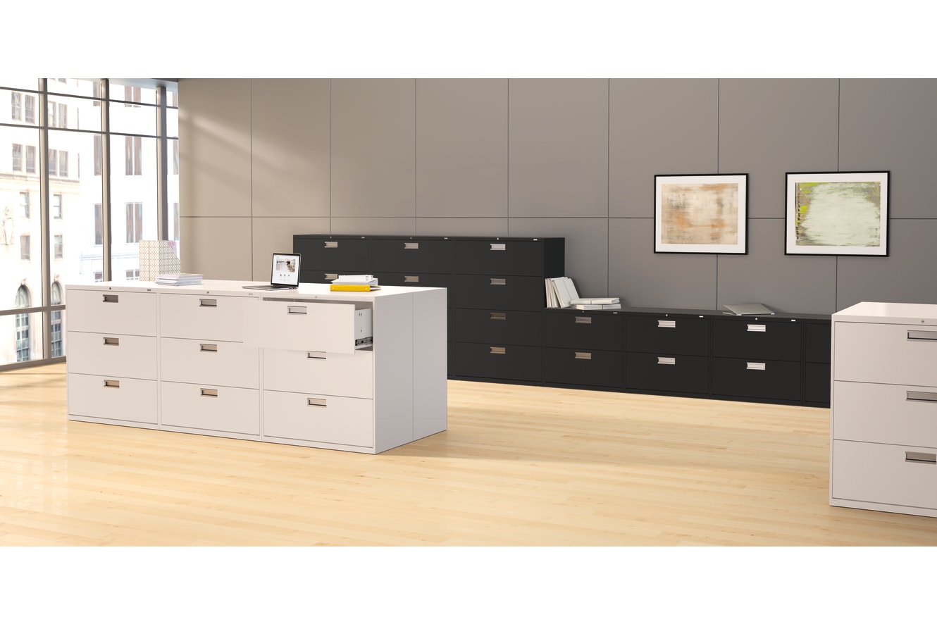 The image depicts a modern office space with a white filing cabinet and a black storage unit in the foreground, set against a backdrop of large windows and framed artwork on the walls.