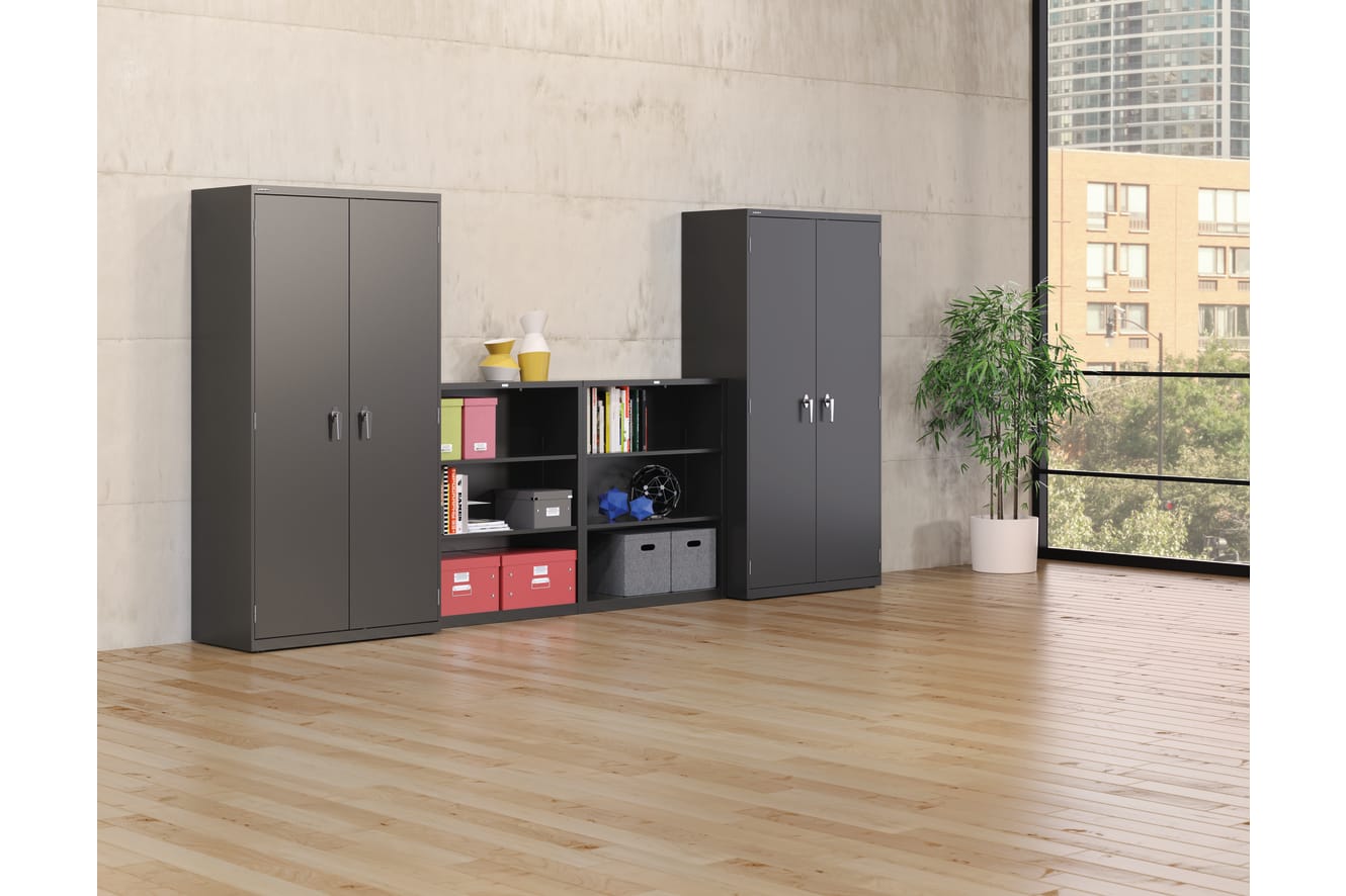 The image depicts a modern and minimalist office or home interior, featuring a set of black storage cabinets with shelves and compartments, arranged against a plain concrete wall. The floor is made of light-colored hardwood, and a potted plant adds a touc