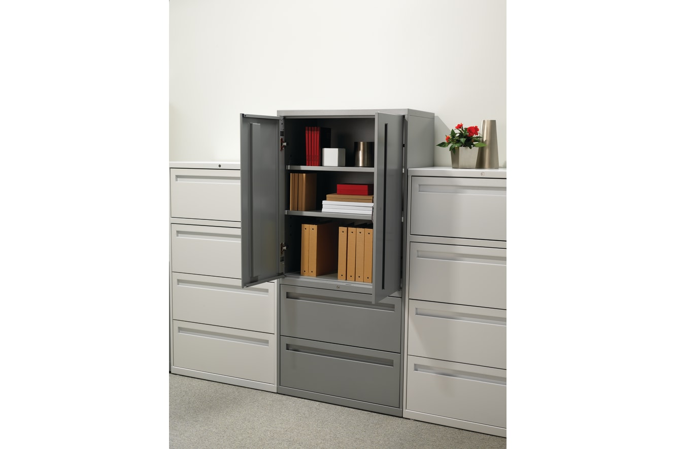 A gray metal storage cabinet with multiple drawers and shelves, containing various office supplies and documents, stands in a clean, minimalist office environment.