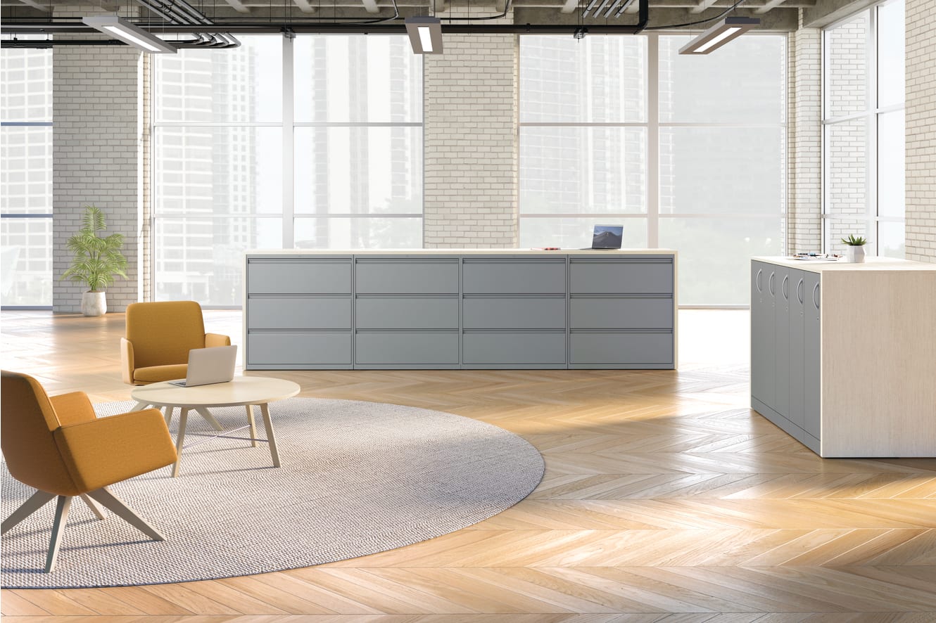 A modern, minimalist office space with gray storage units, yellow chairs, and a round rug on a hardwood floor, surrounded by large windows letting in natural light.