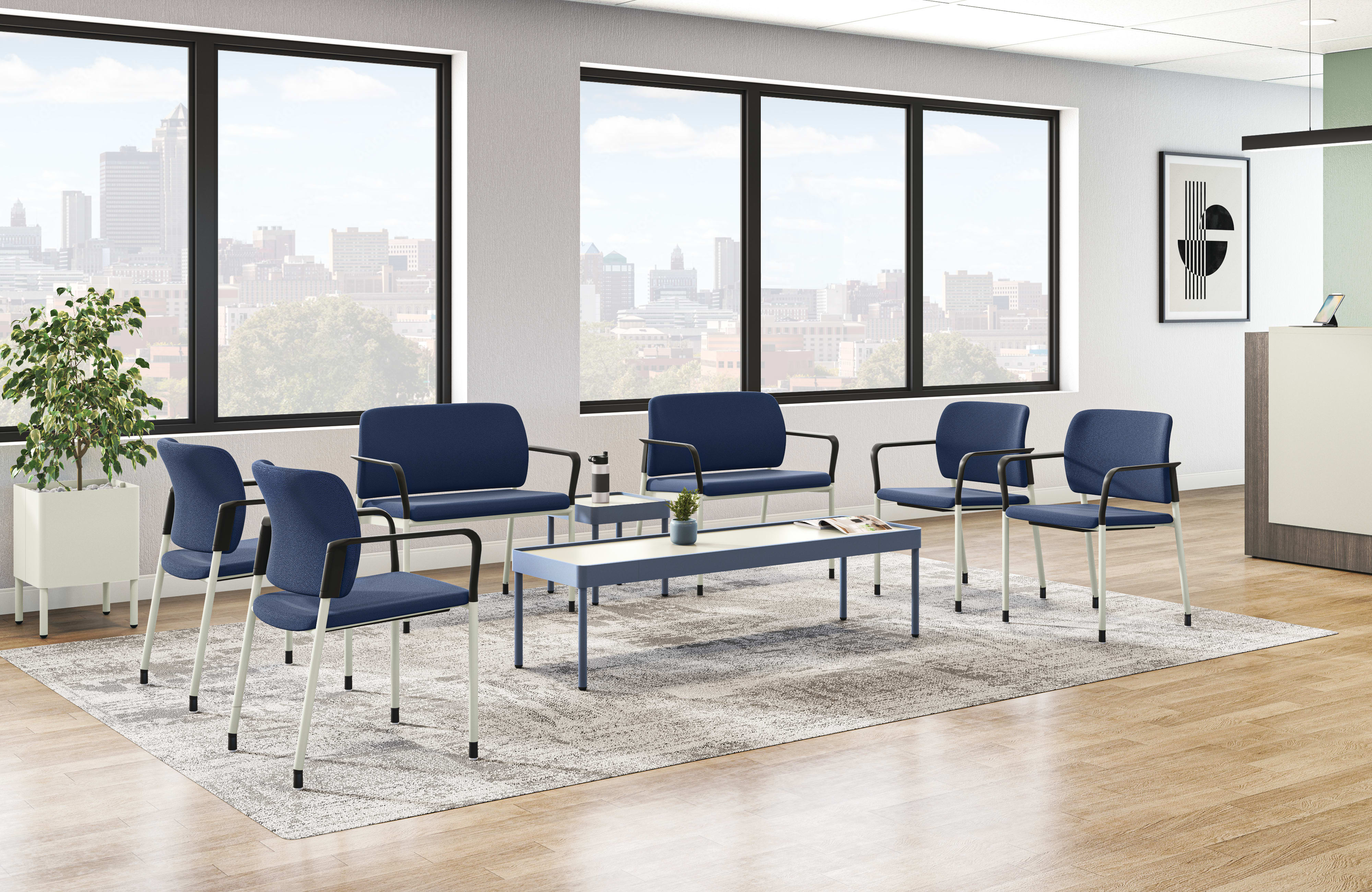 Modern office lounge with blue upholstered chairs, matching coffee table, large windows with city view, indoor plant, and framed wall art on light gray walls.