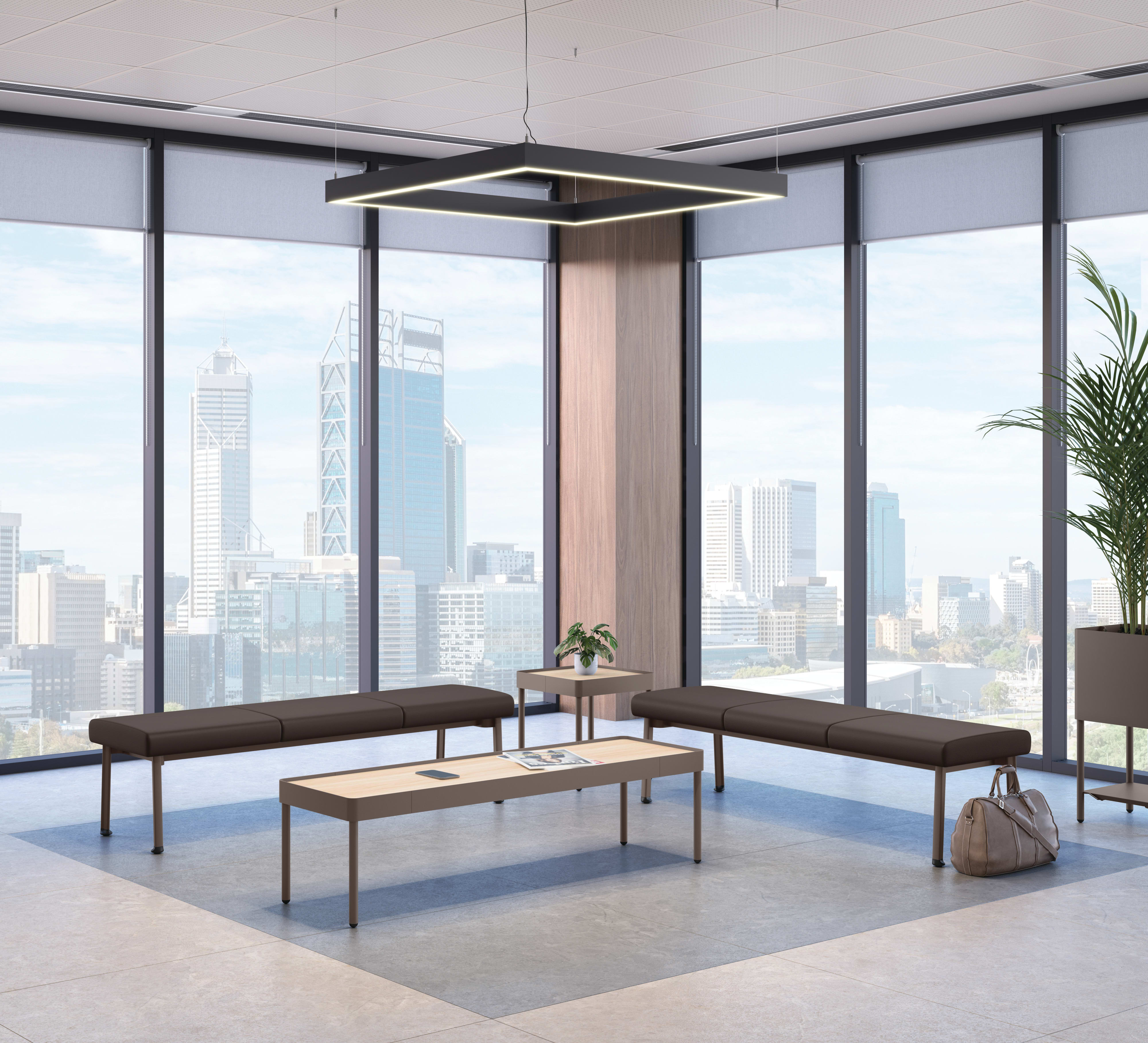 Modern office lounge with two long cushioned benches and a rectangular coffee table, set against floor-to-ceiling windows showcasing a city skyline, accented by a geometric ceiling light and potted plant.