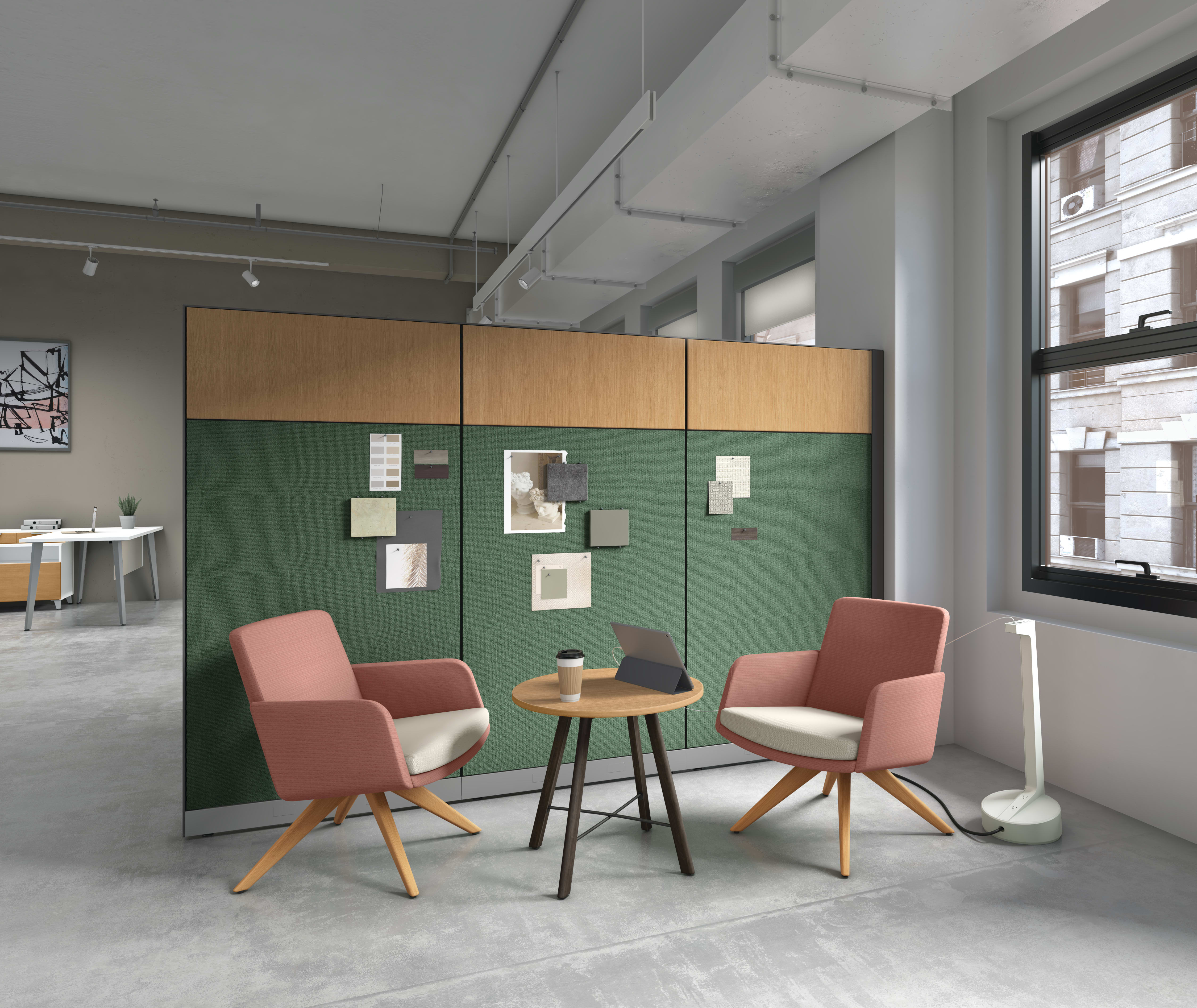 Office seating arrangement with two pink armchairs, round wooden table, and green partition wall with notes.