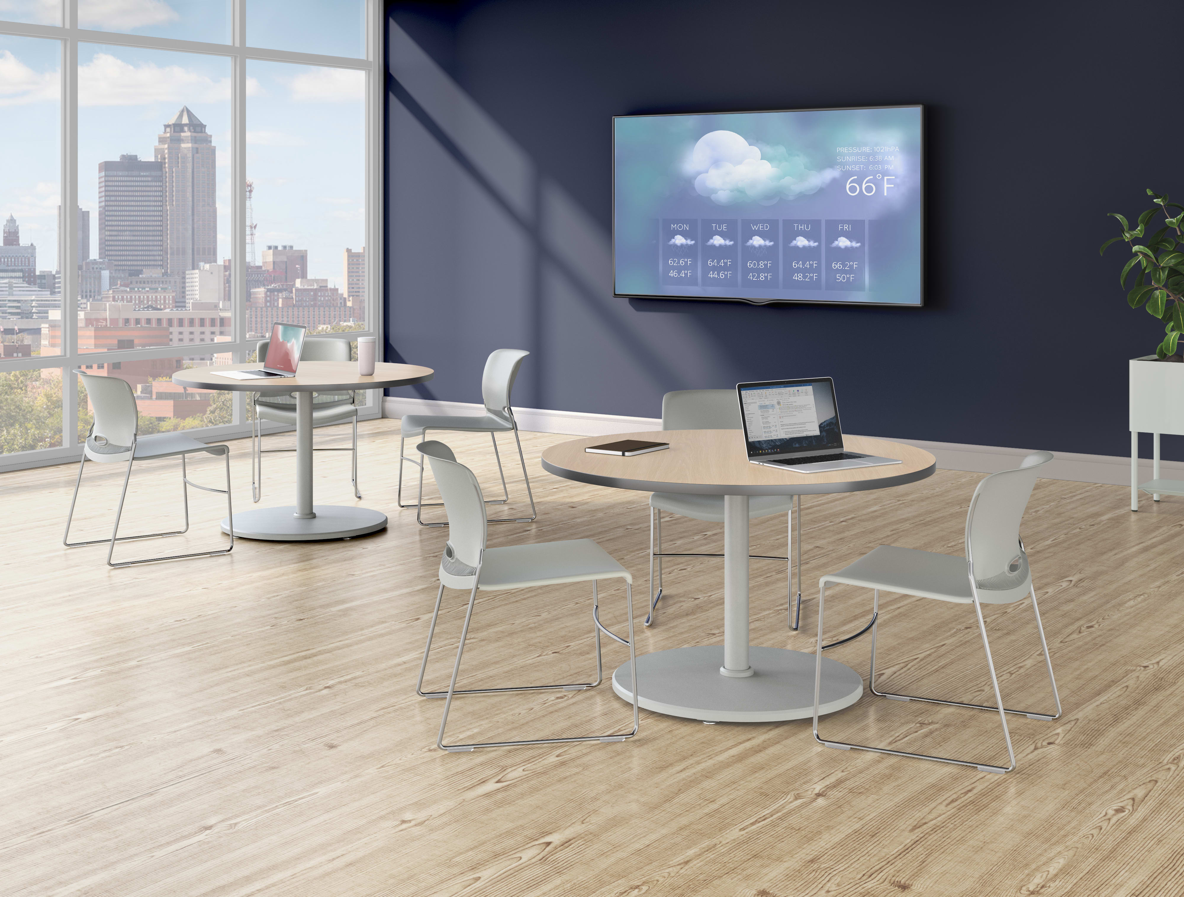 A bright lounge area with round tables, light-colored chairs, a wall-mounted display showing weather information, and large windows overlooking a city skyline.