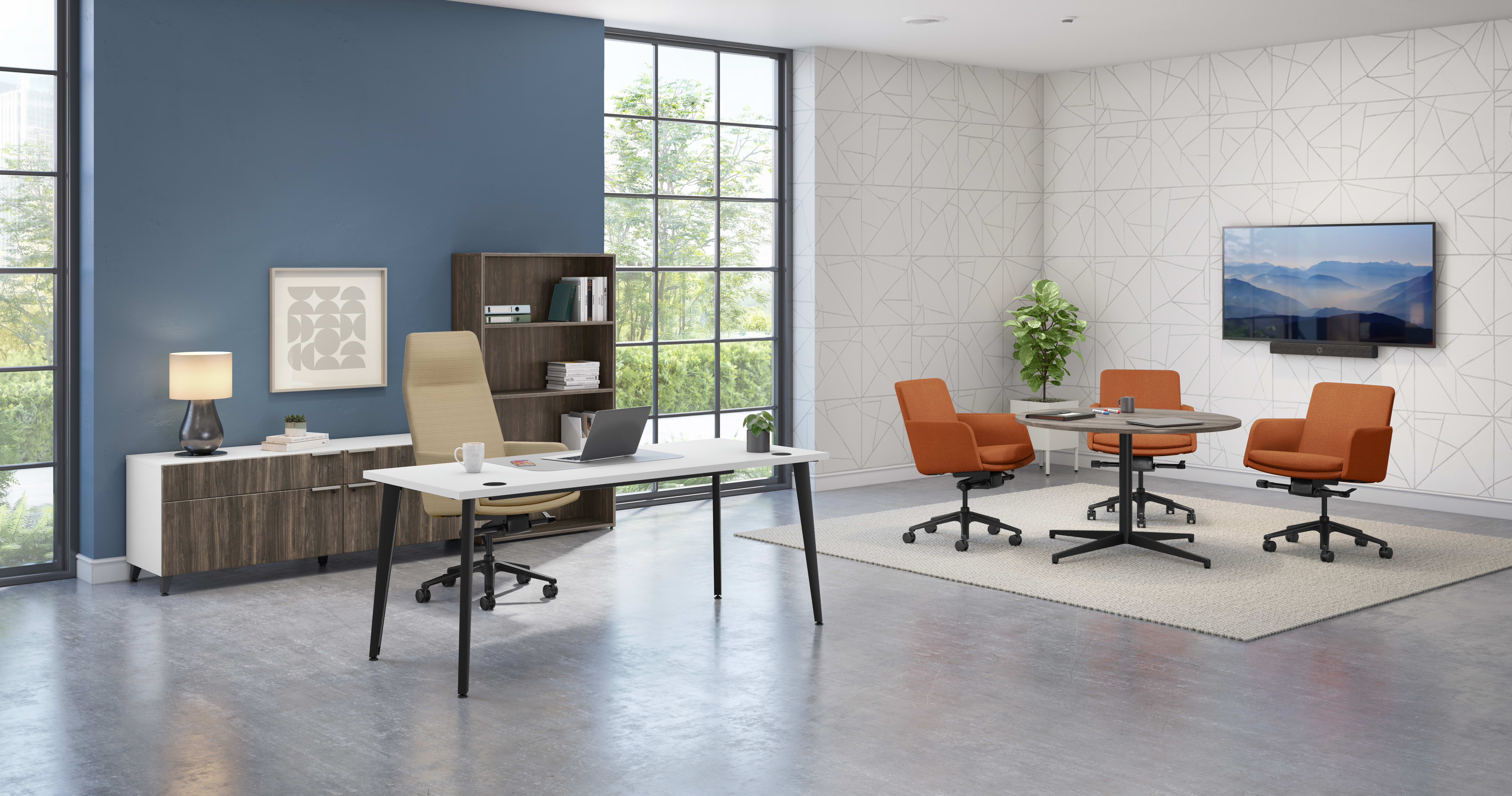 Modern office setting with a white desk and beige chair on the left, wood credenza and bookcase against a blue accent wall, and a meeting area on the right featuring four orange chairs around round tables on a neutral rug, with large windows and geometric