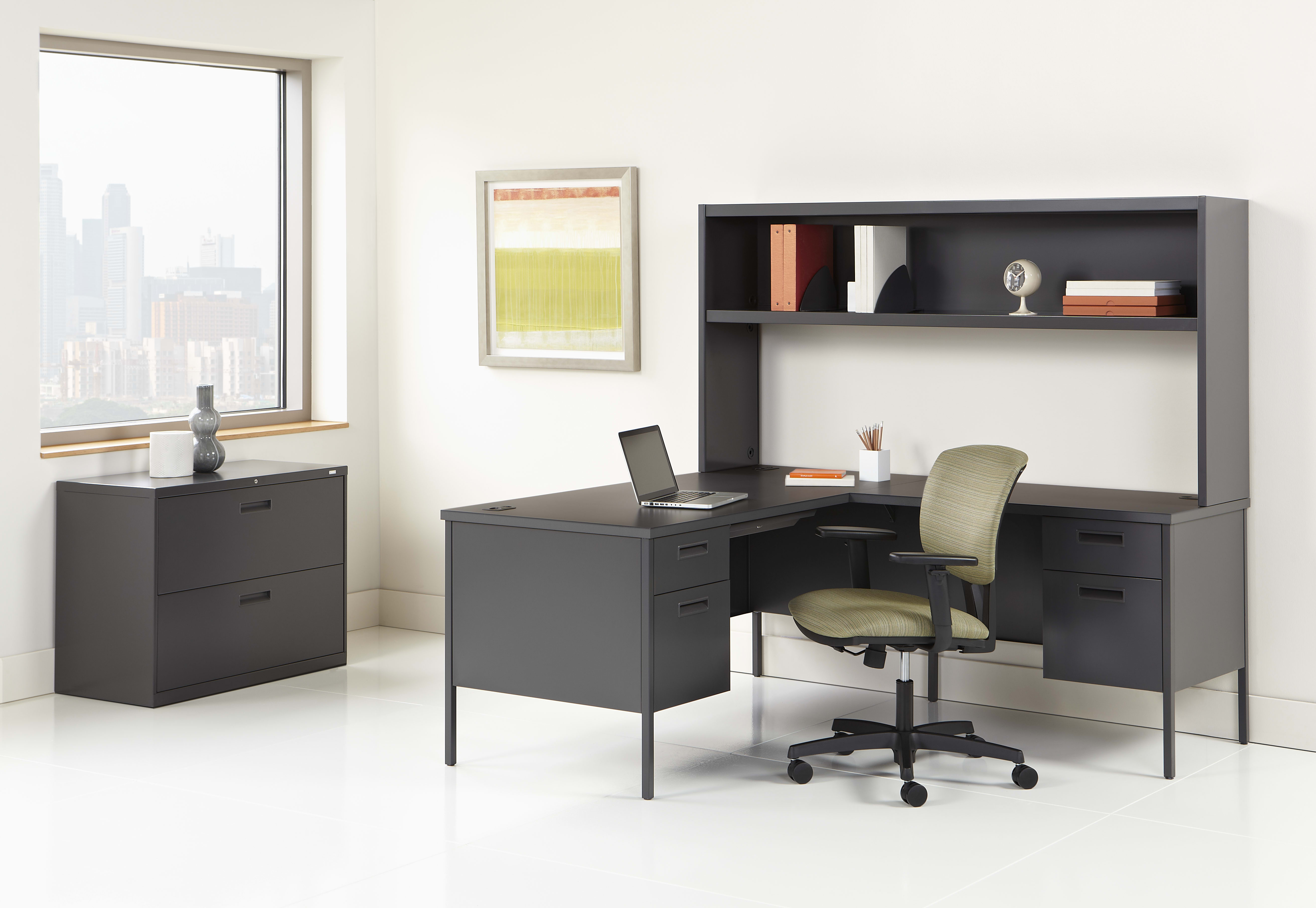 A modern and minimalist home office setup with a black desk, shelving unit, and a green office chair against a white wall with a large window.