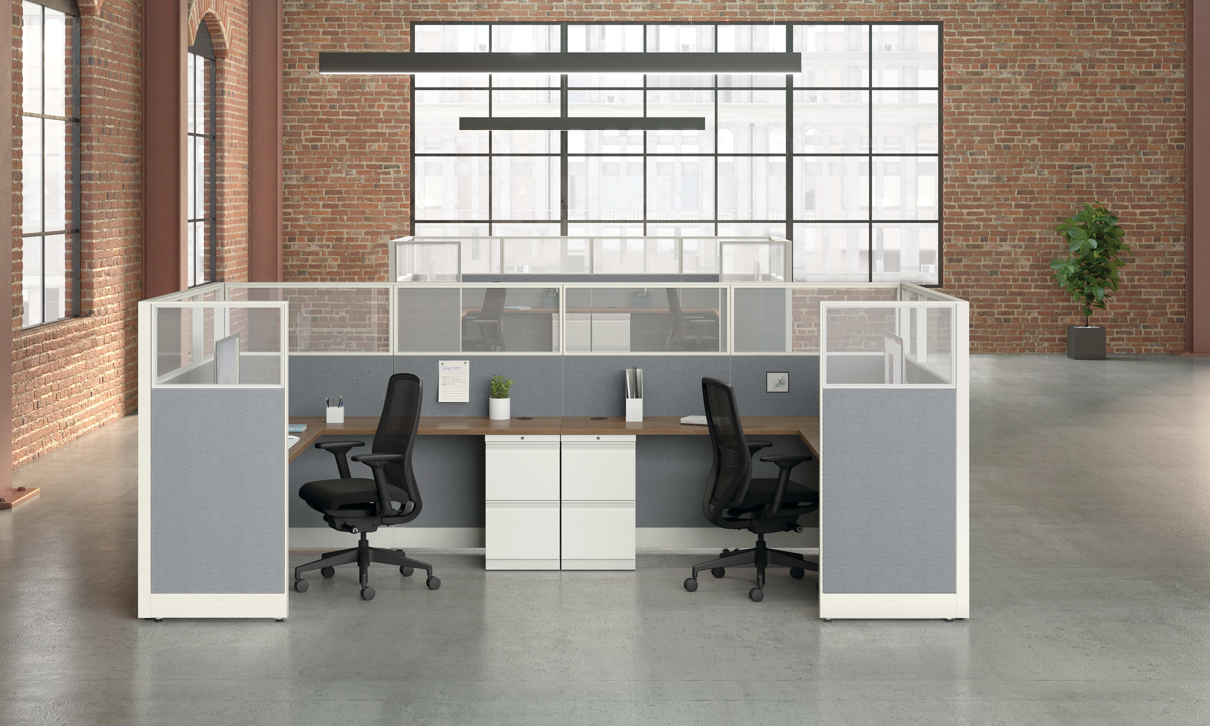 A modern, minimalist office space with white and gray cubicles, chairs, and a potted plant against a brick wall background.