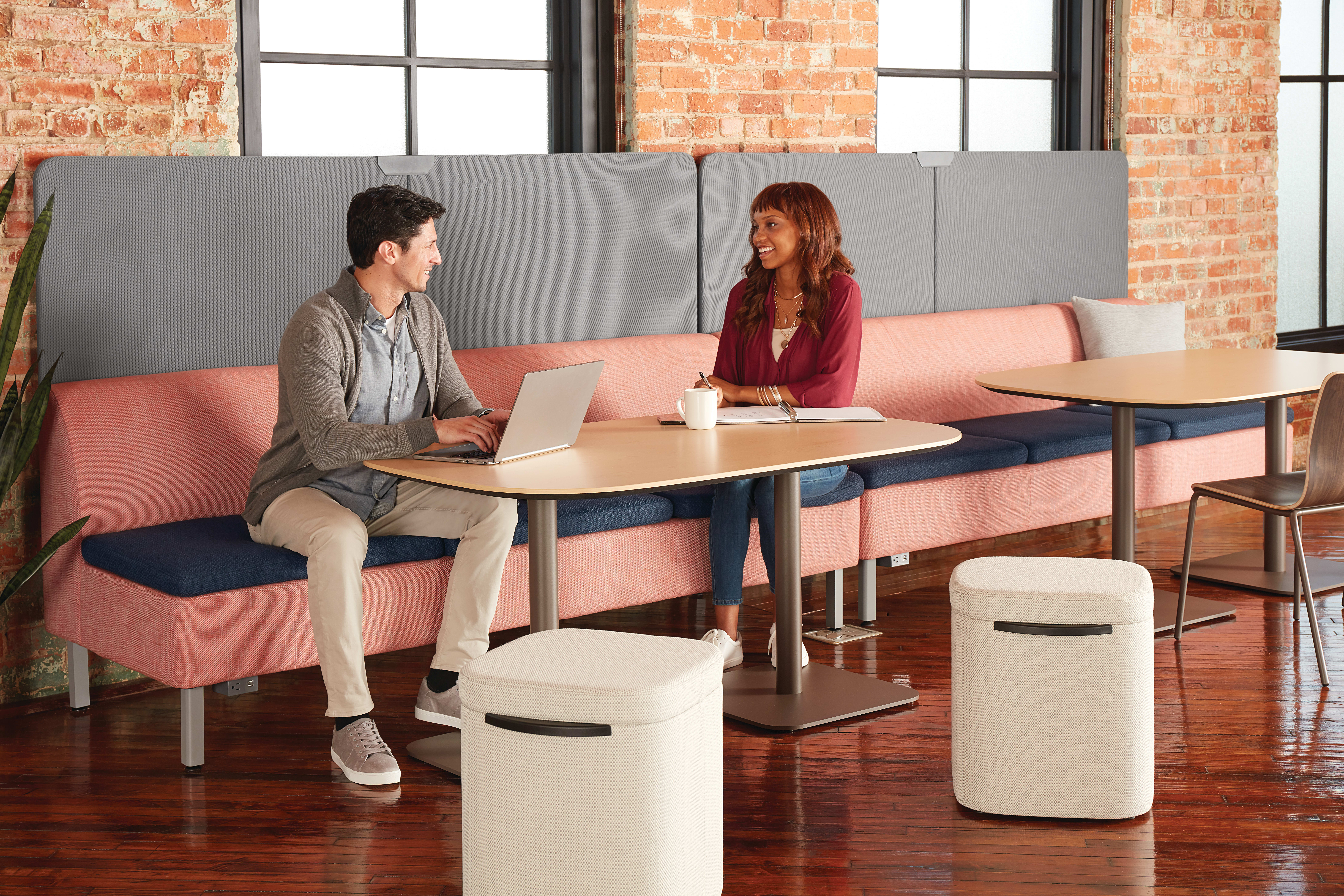 Two people, a man and a woman, are seated at a table in a modern office-like setting with brick walls and large windows in the background.