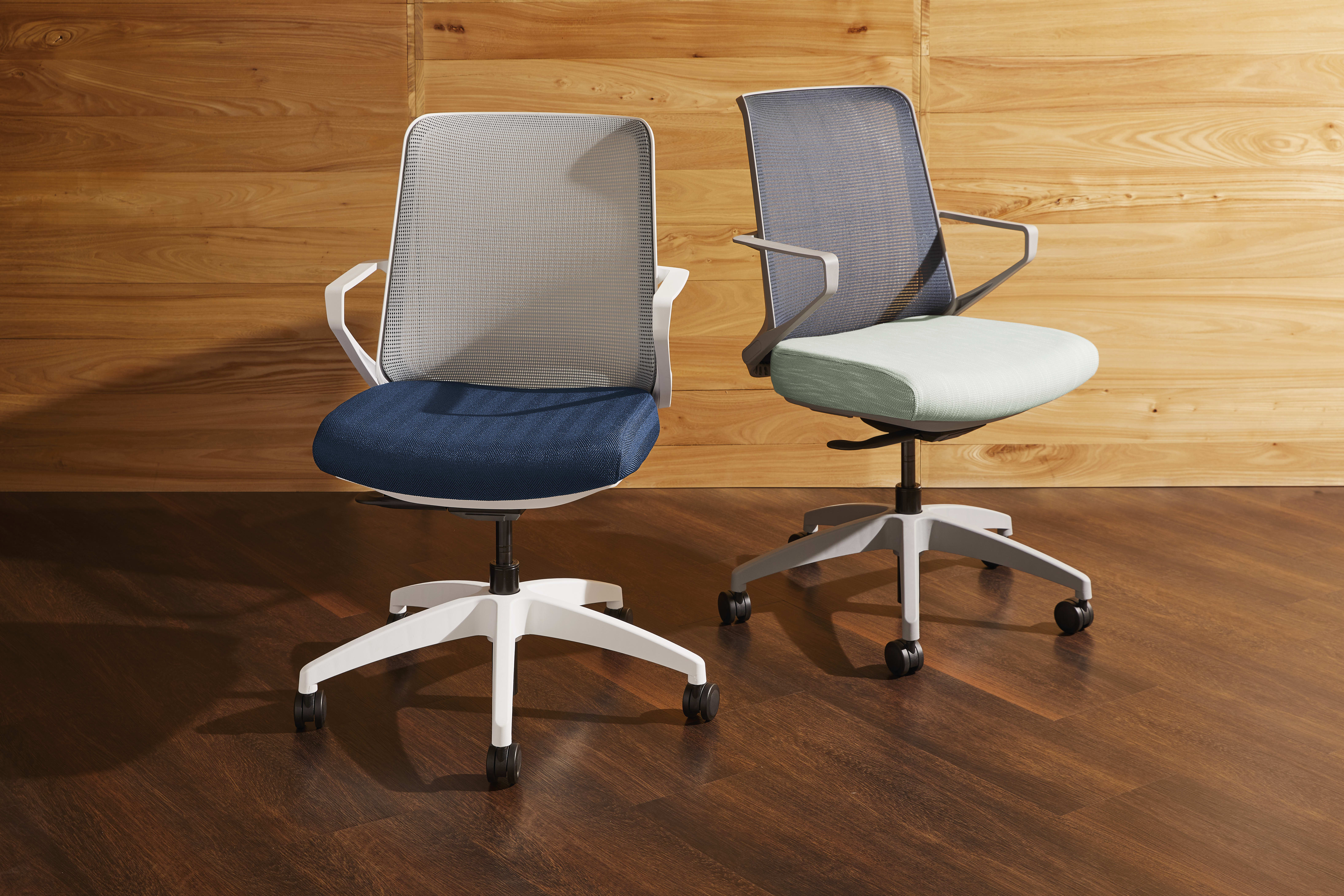 Two modern office chairs with mesh backs and padded seats sit on a wooden floor against a wooden wall background.