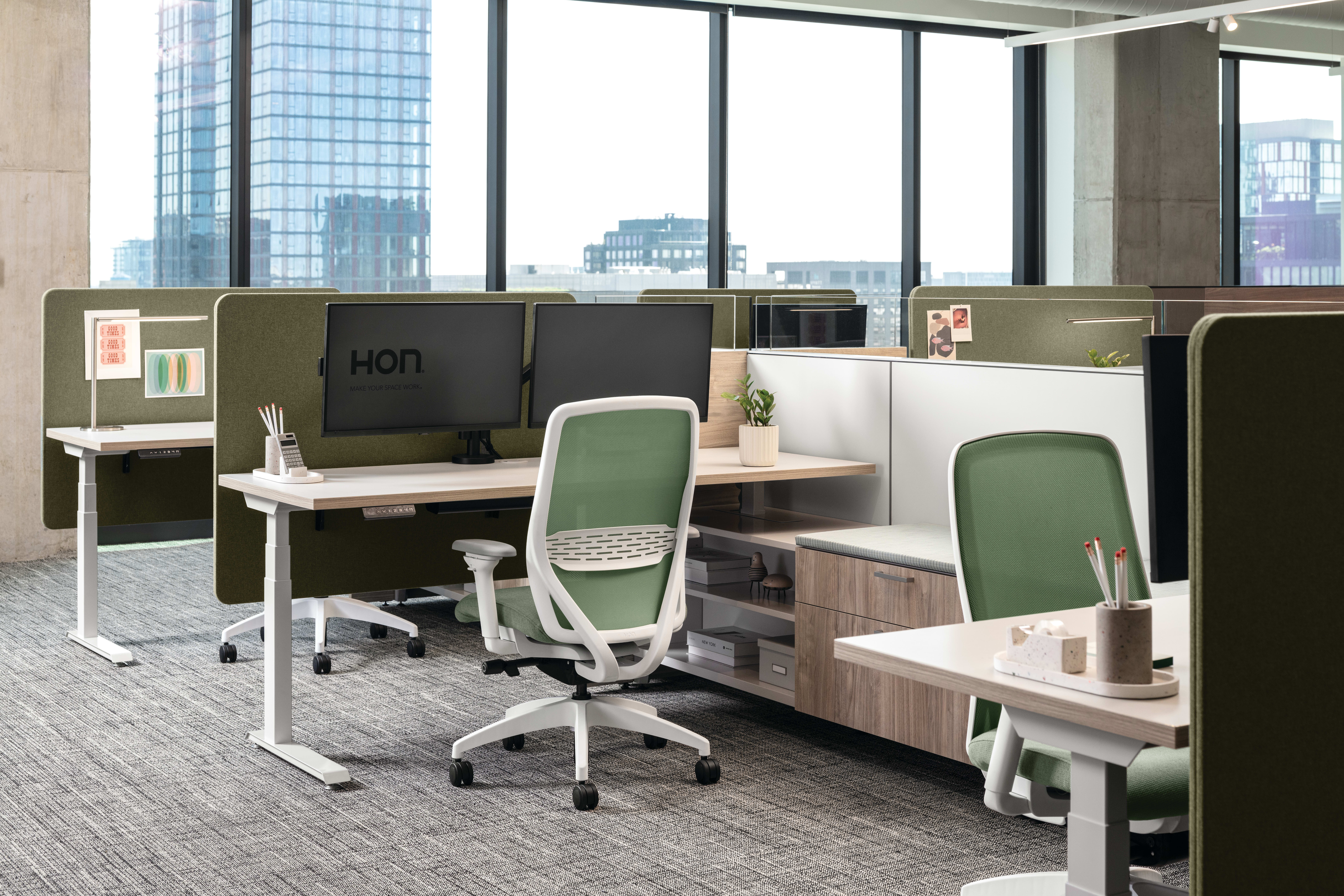 Open workstation cluster with height-adjustable desks, upholstered privacy panels, dual monitor setups, ergonomic task chairs, and integrated storage, complemented by bench seating with cushion top, personal lighting, and accessory trays.