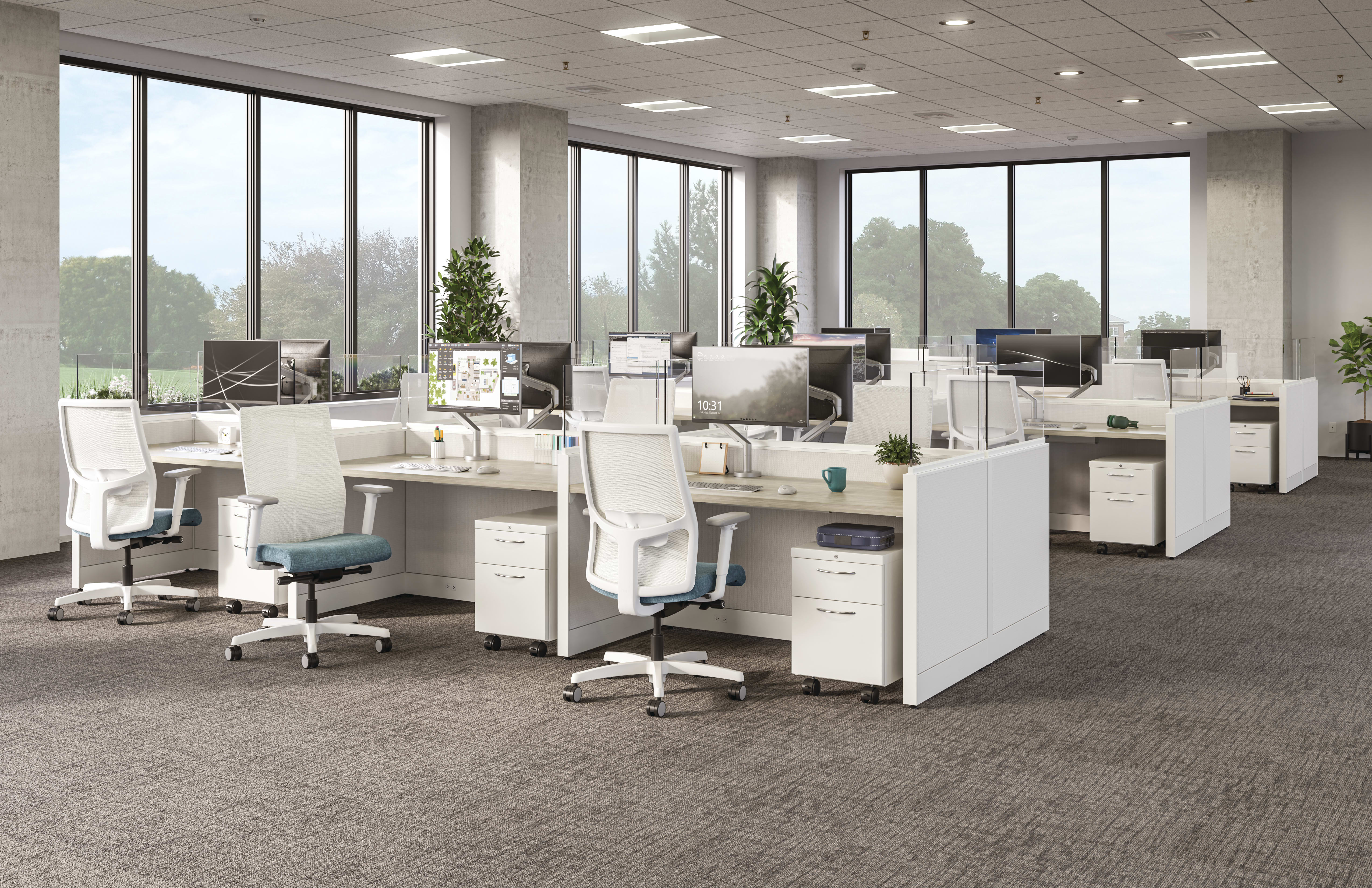 Open office layout with white workstations, ergonomic chairs, desktop monitors, and large windows overlooking greenery.