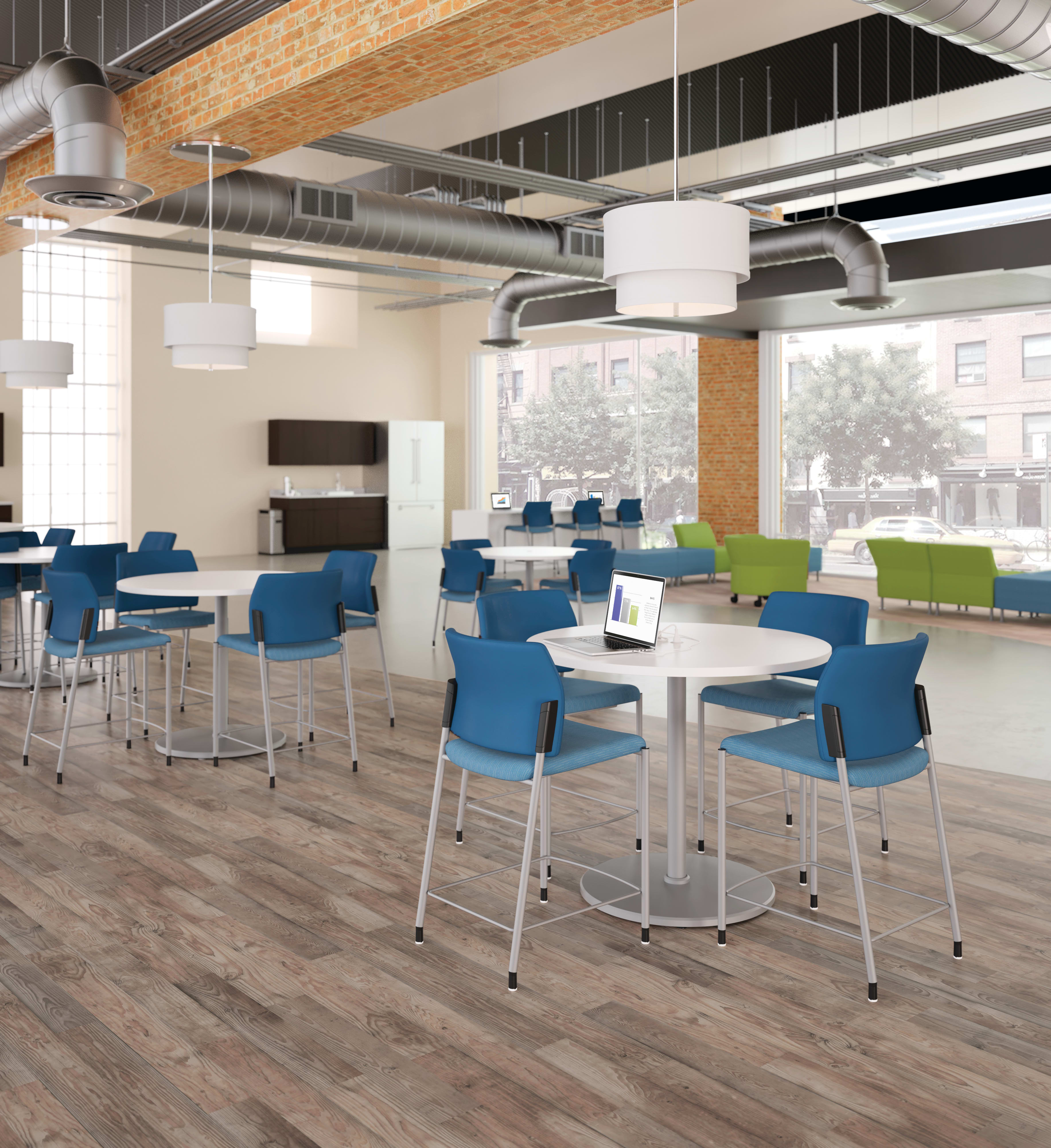 A modern and open office space with a mix of blue and green chairs, round tables, and exposed ceiling ducts, creating a bright and collaborative work environment.