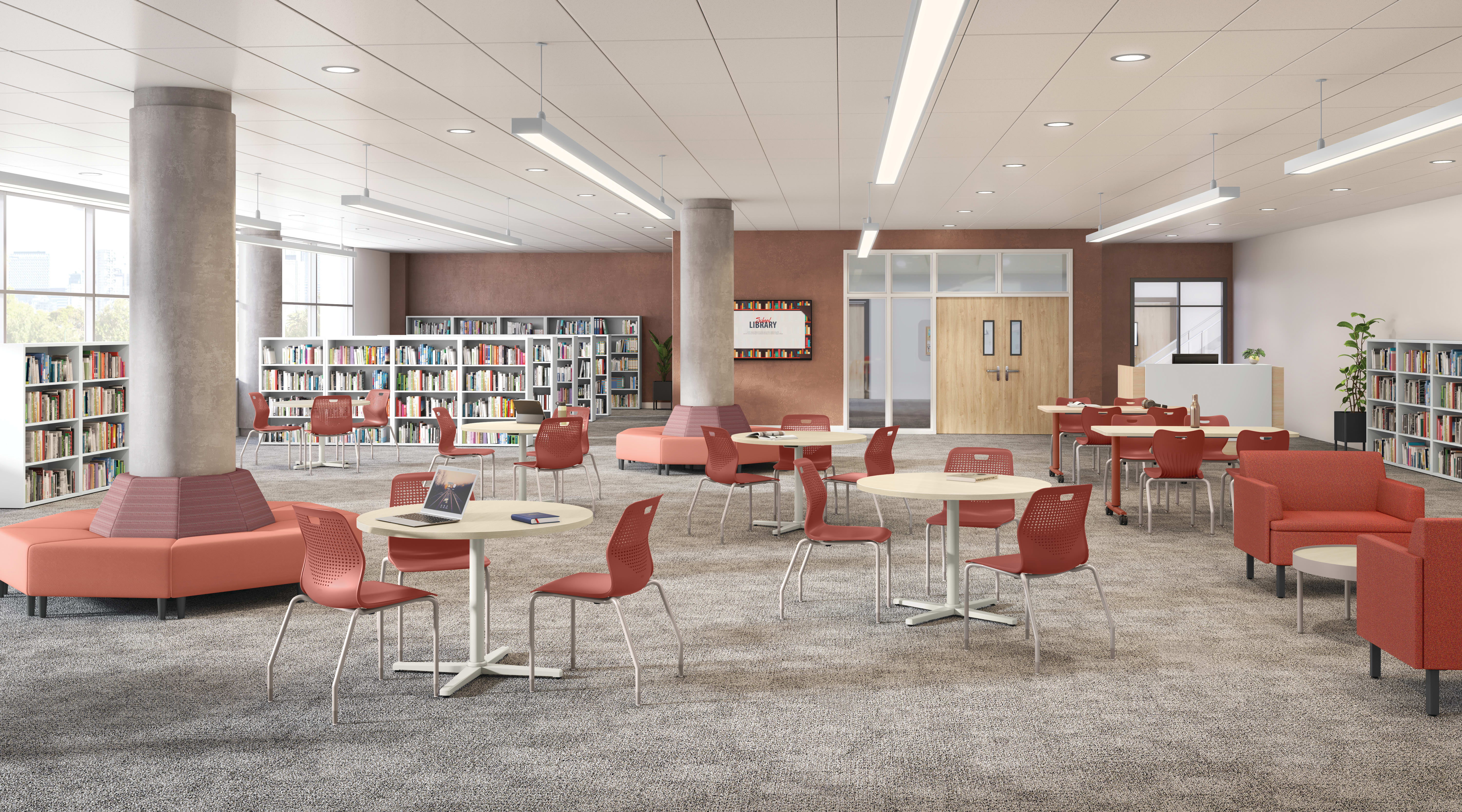 Modern library commons with bookshelves, round study tables, red chairs, lounge seating, structural columns, carpeted flooring, and bright overhead lighting in an open interior space.