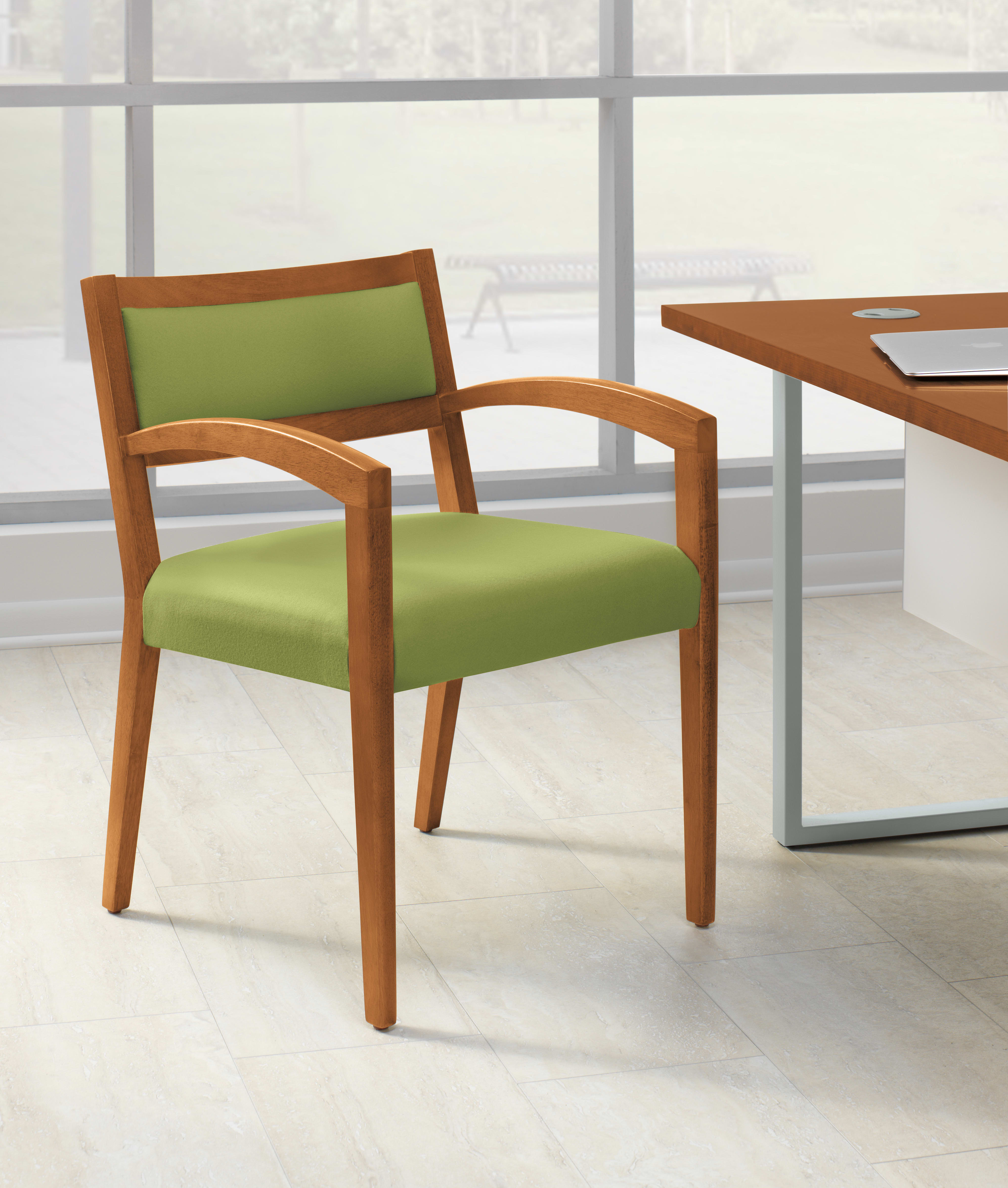 A wooden chair with a green cushioned seat sits in a bright, minimalist office environment with a large window in the background.