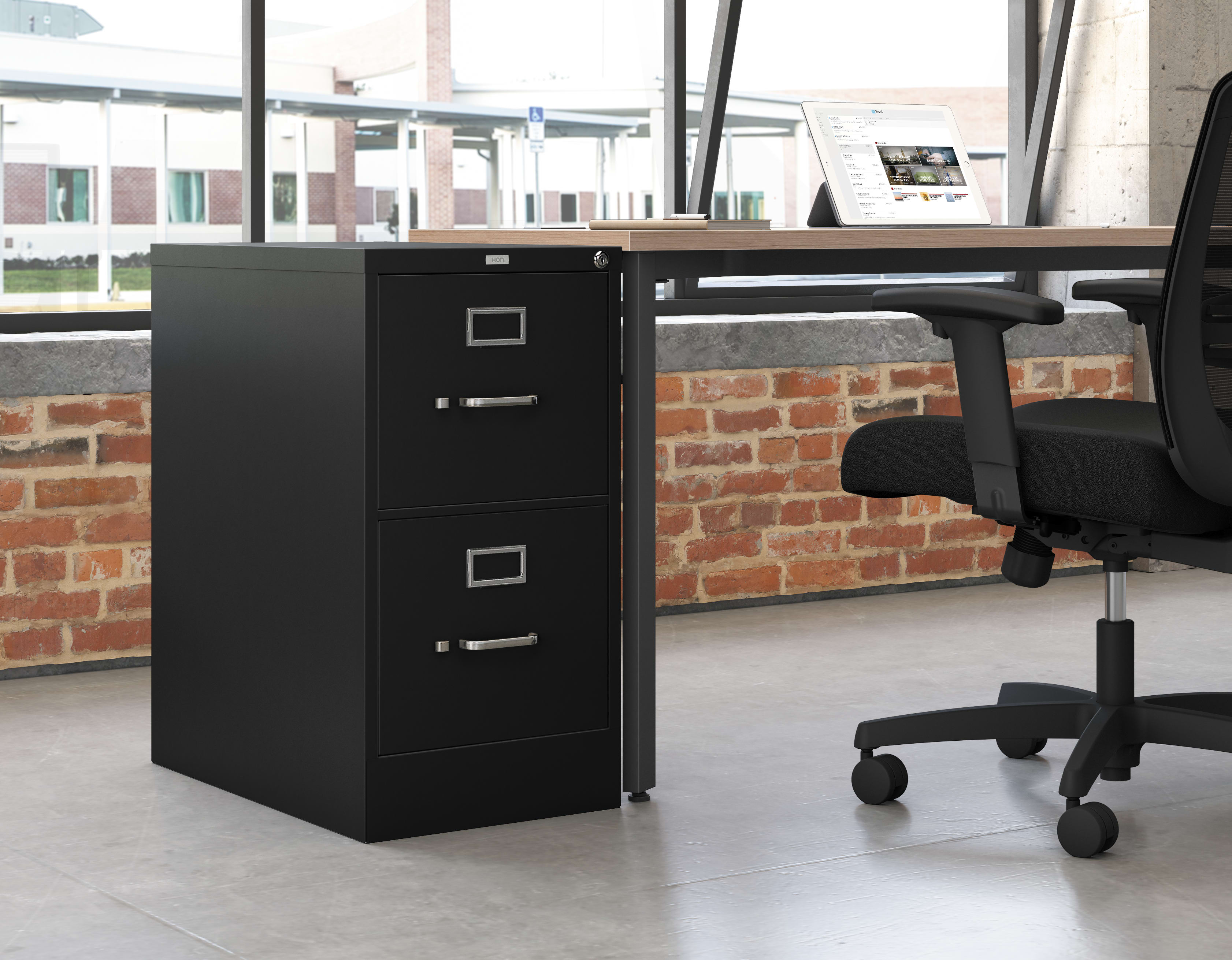 A black filing cabinet stands in the foreground, with a desk and office chair visible in the background, set against a brick wall.