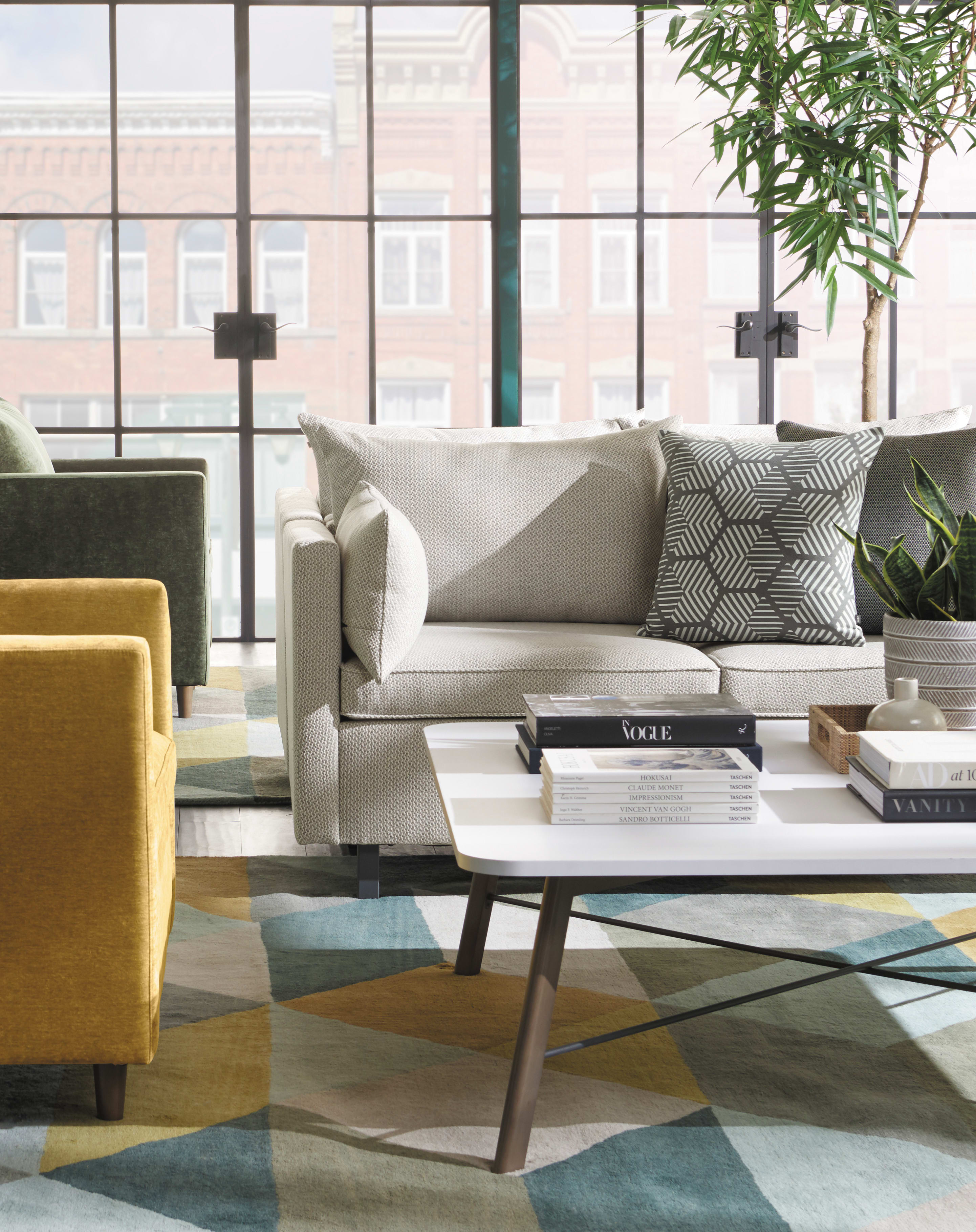 A cozy and modern living room with a beige sofa, patterned throw pillows, a yellow armchair, and a glass coffee table on a colorful area rug, set against a backdrop of large windows overlooking a city skyline.