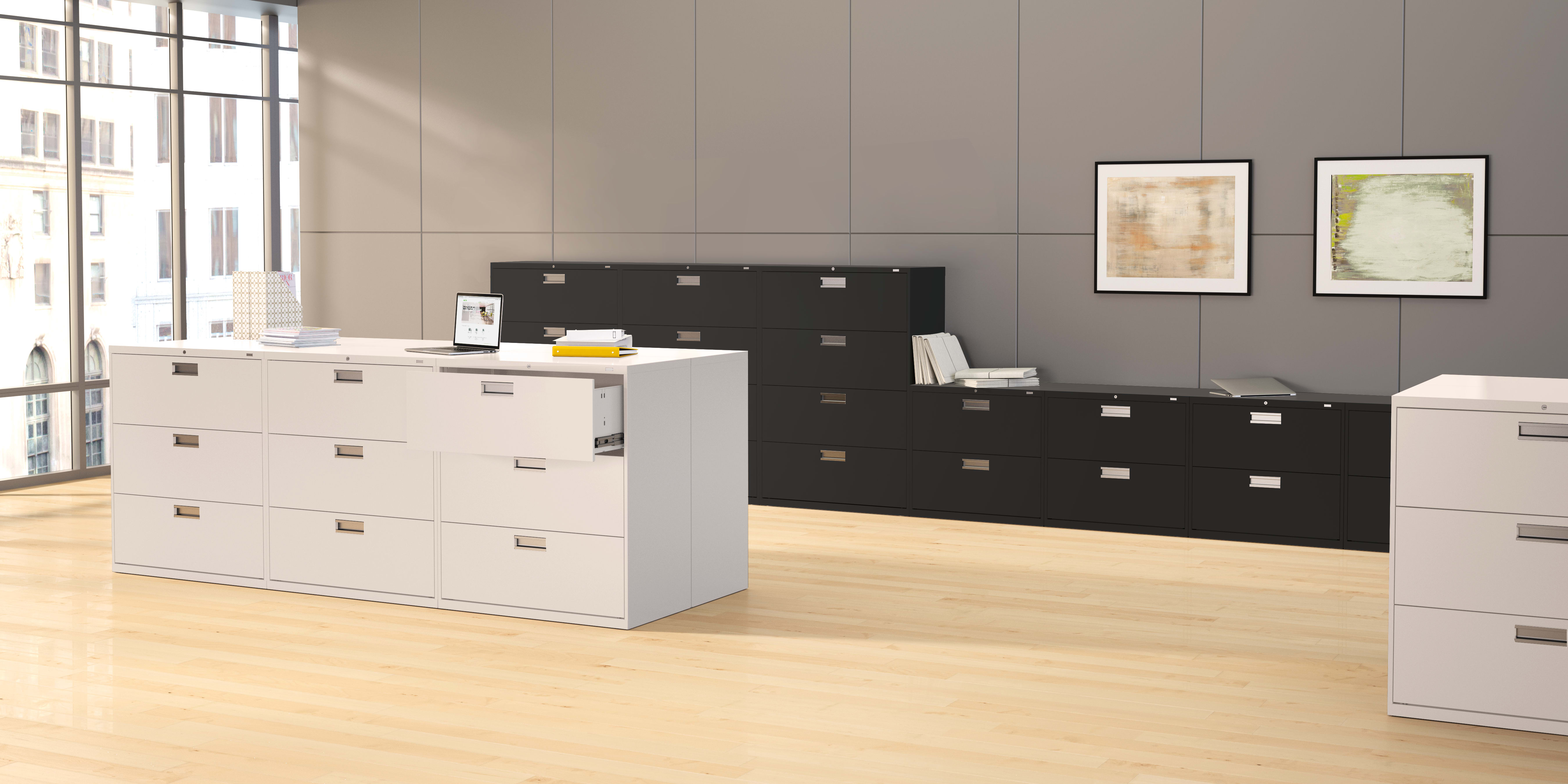 The image depicts a modern office space with a white filing cabinet and a black storage unit in the foreground, set against a backdrop of large windows and framed artwork on the walls.
