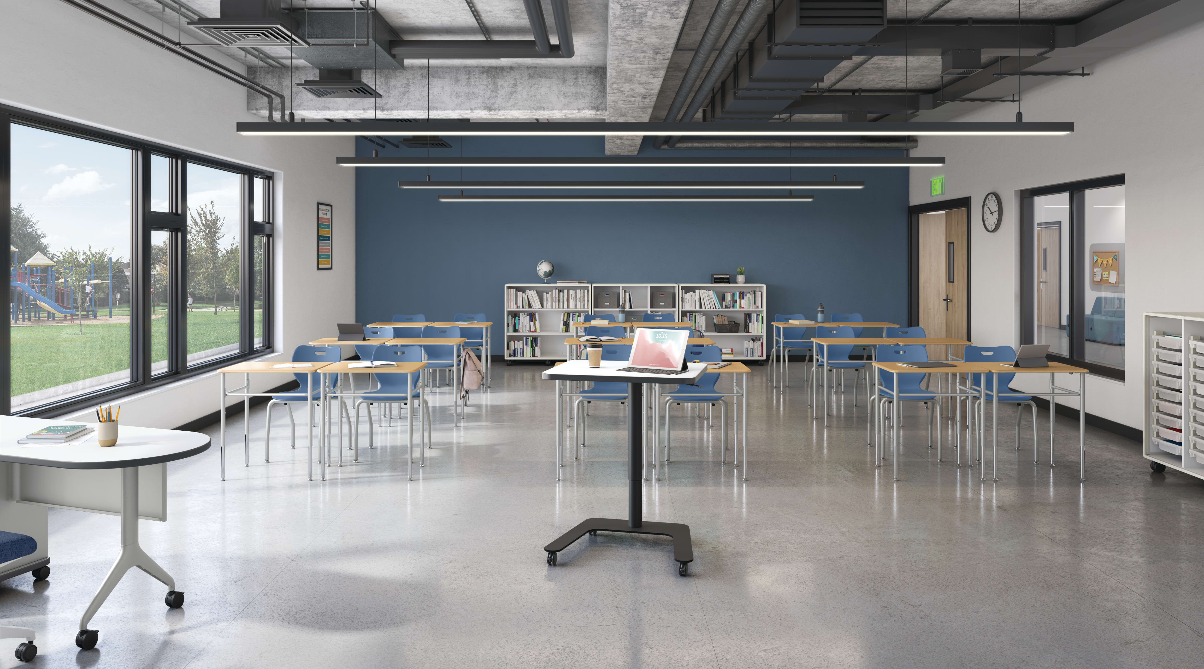 Modern classroom with flexible desks and chairs, large windows, blue accent wall, ceiling lighting, bookshelves, and teacher workstation with laptop and coffee cup.