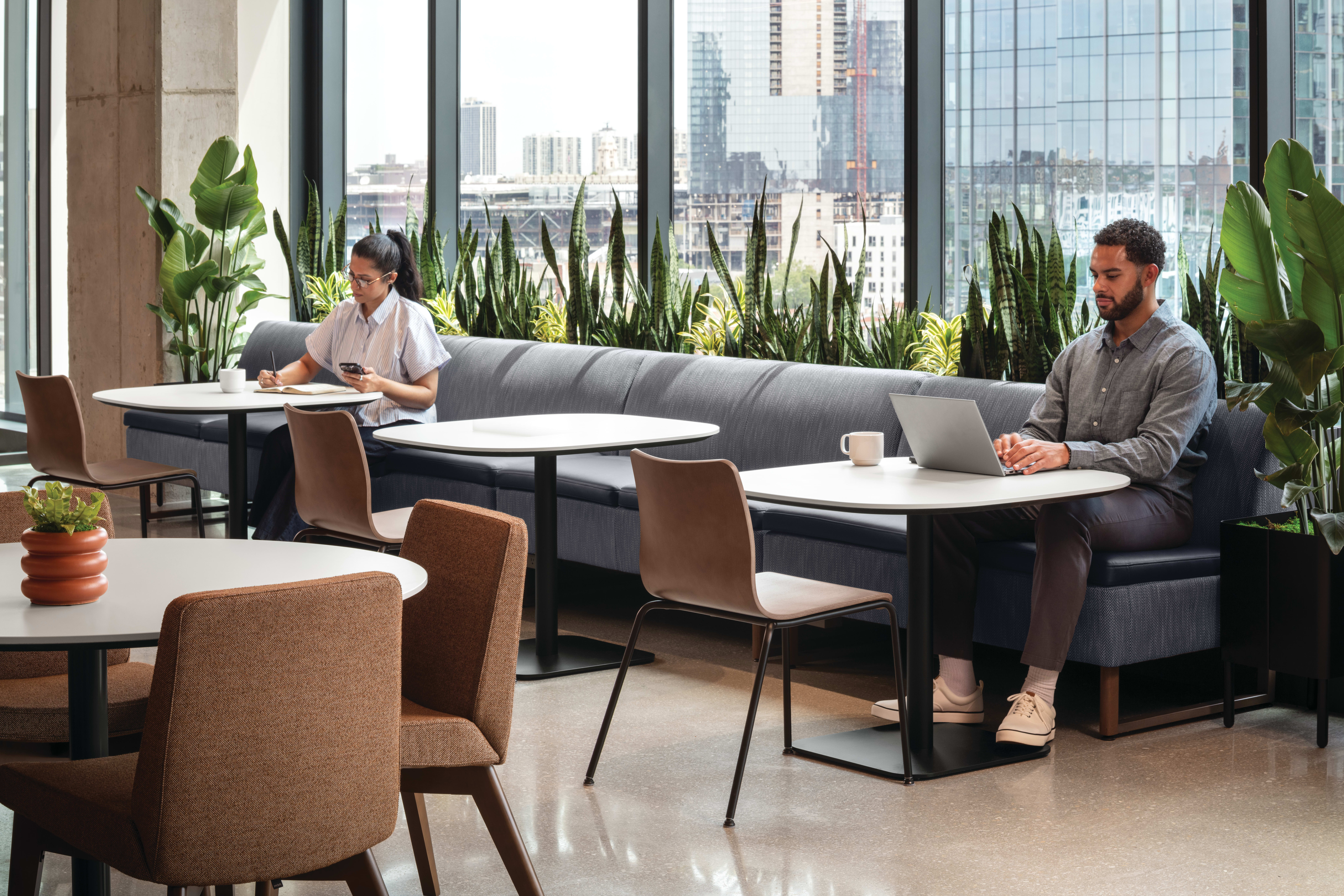 Café-style seating area with upholstered banquette, round pedestal tables, and mixed-material guest chairs, complemented by integrated planter with greenery and adjacent lounge seating.