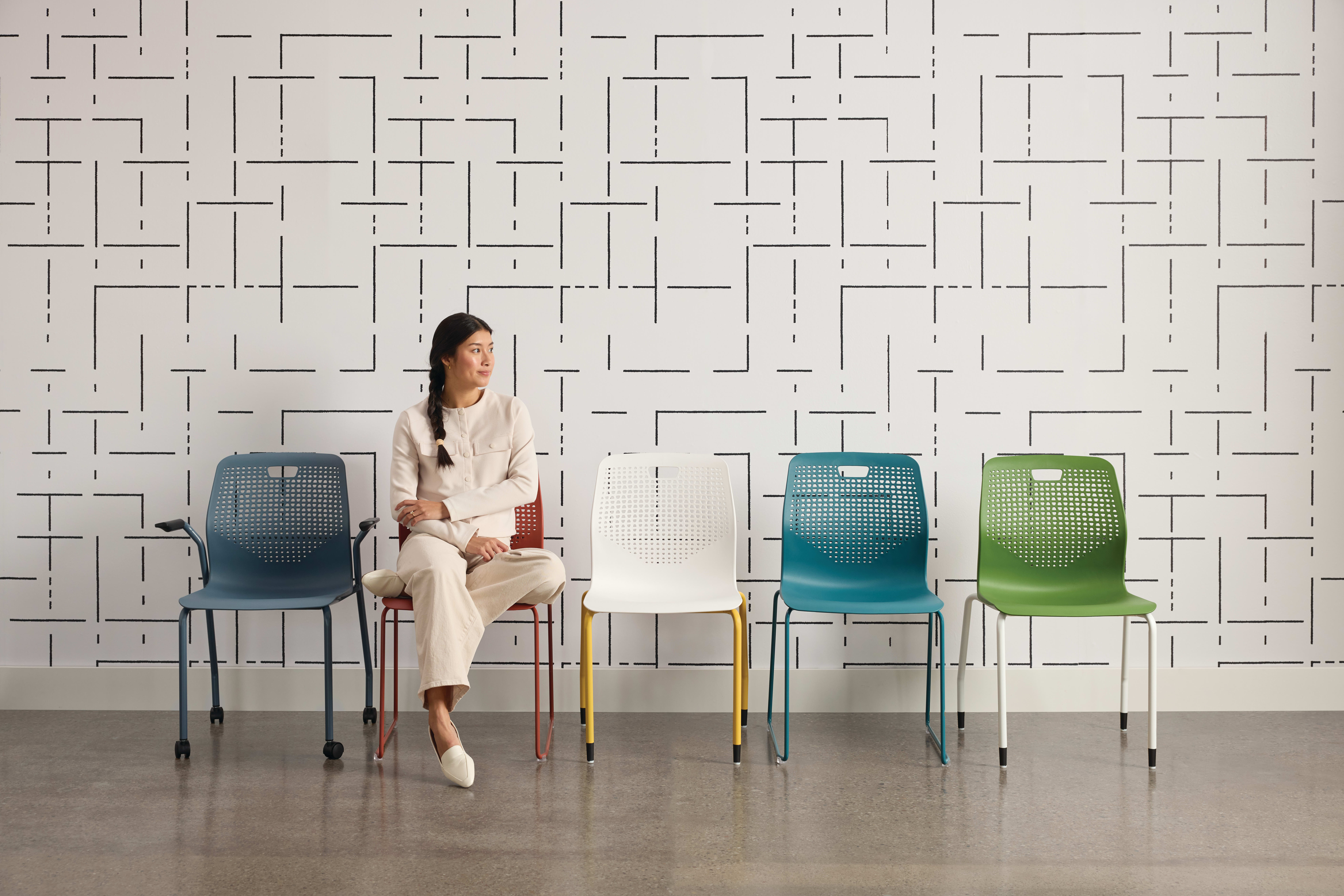 A woman in a beige outfit sits on a chair in front of a patterned wall, surrounded by colorful chairs in various shades of blue and green.