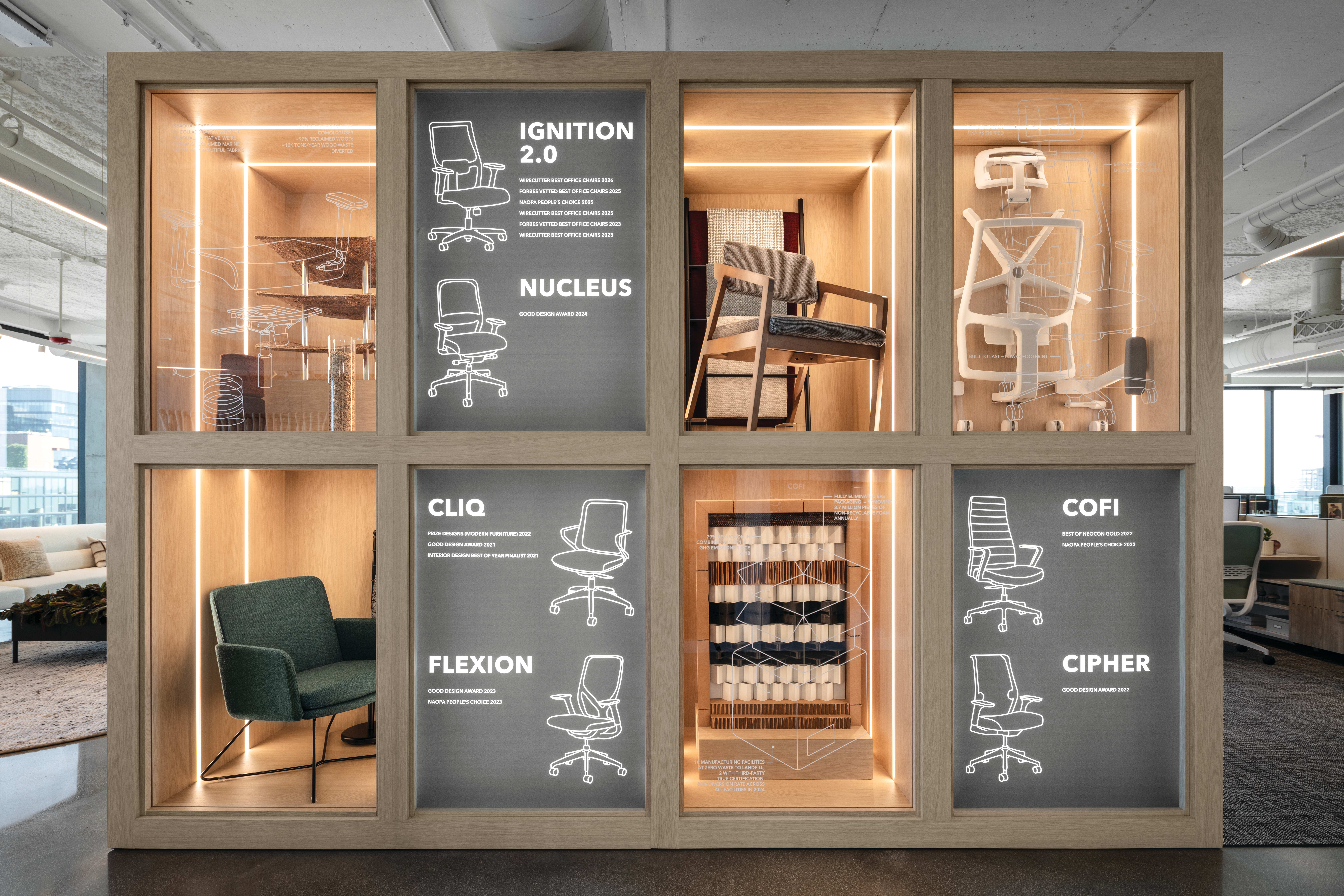 Showcase wall with illuminated wood display niches featuring multiple seating designs, including task chairs and a lounge chair, alongside informational panels and material samples.