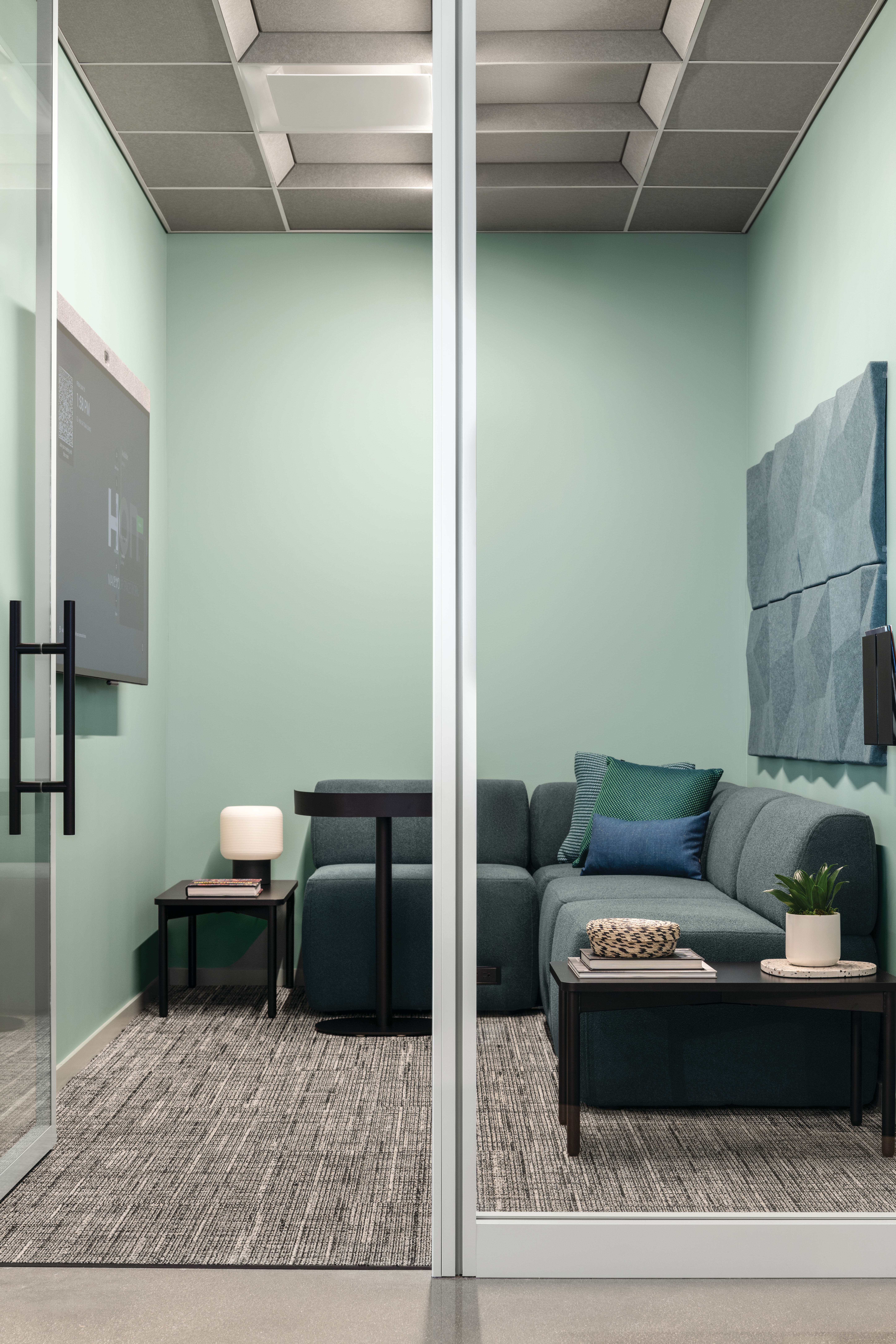 Copilot said: Glass-enclosed focus room with upholstered lounge seating, accent pillows, side tables, integrated media display, acoustic wall panels, and soft lighting.
