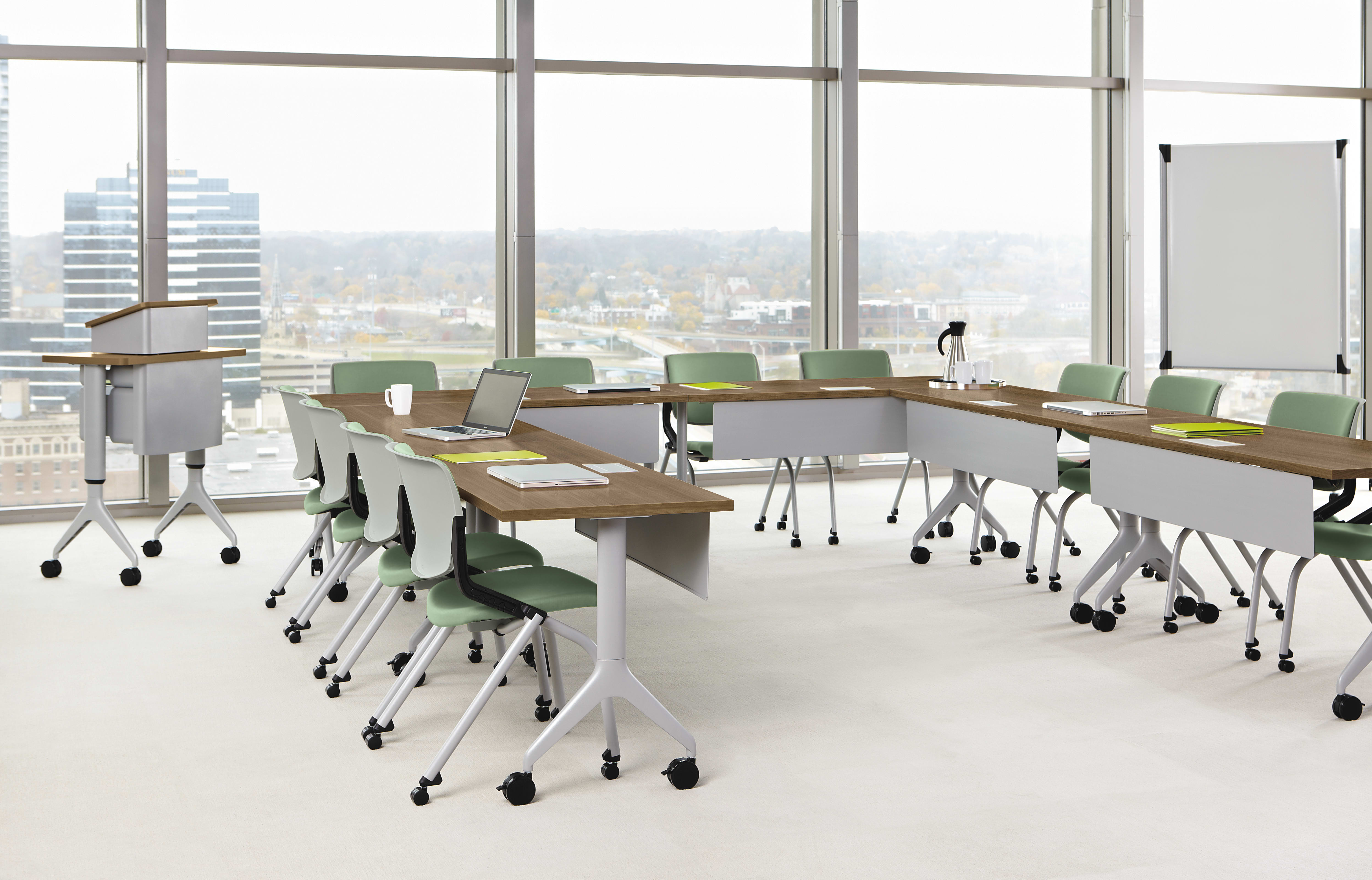 A modern, well-lit classroom or meeting room with large windows overlooking a cityscape, featuring rows of desks and chairs arranged for collaborative work.