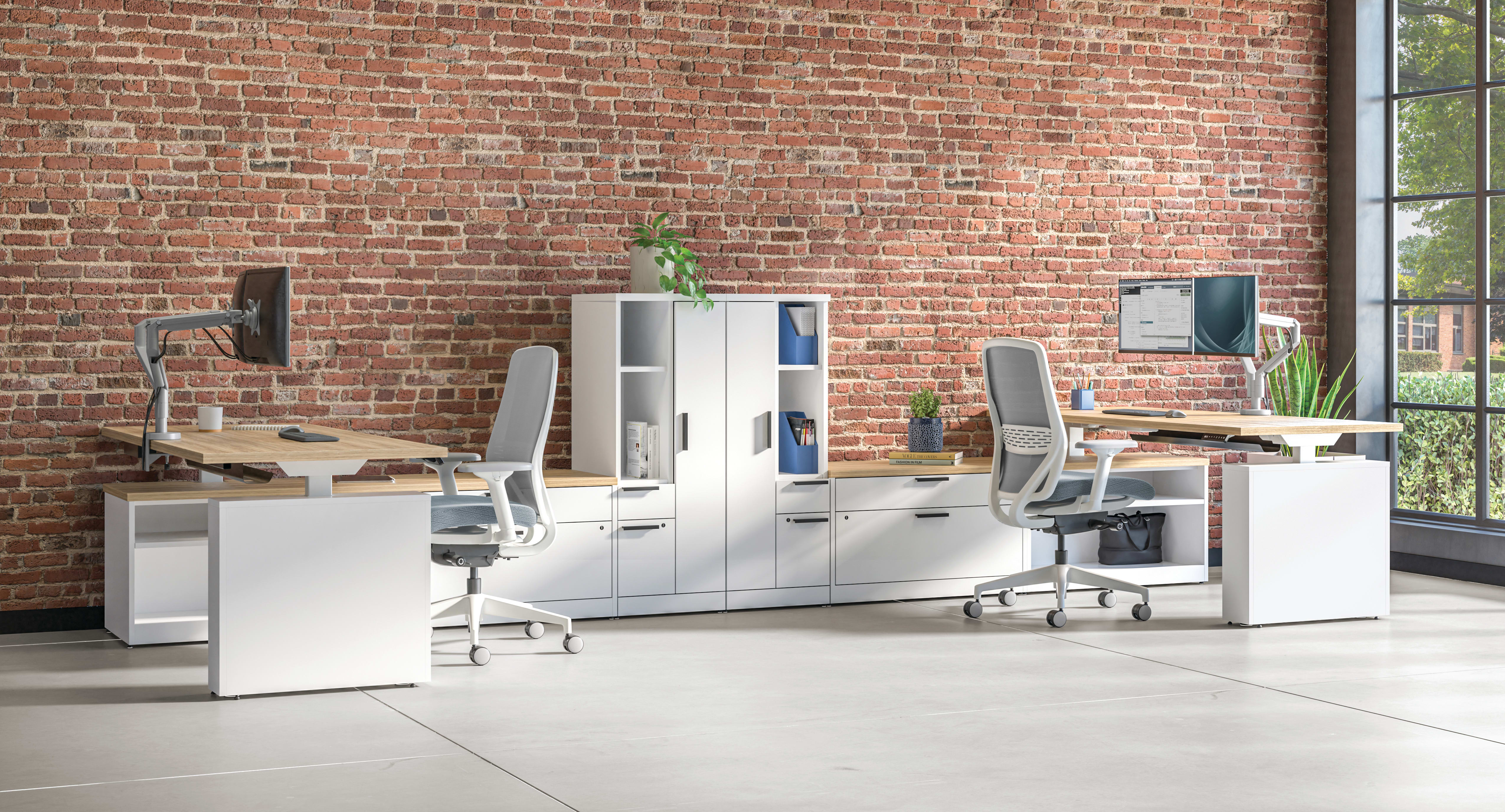 Open office workspace with white desks and ergonomic chairs, modular storage cabinets, computer monitors, and exposed brick wall with large windows.