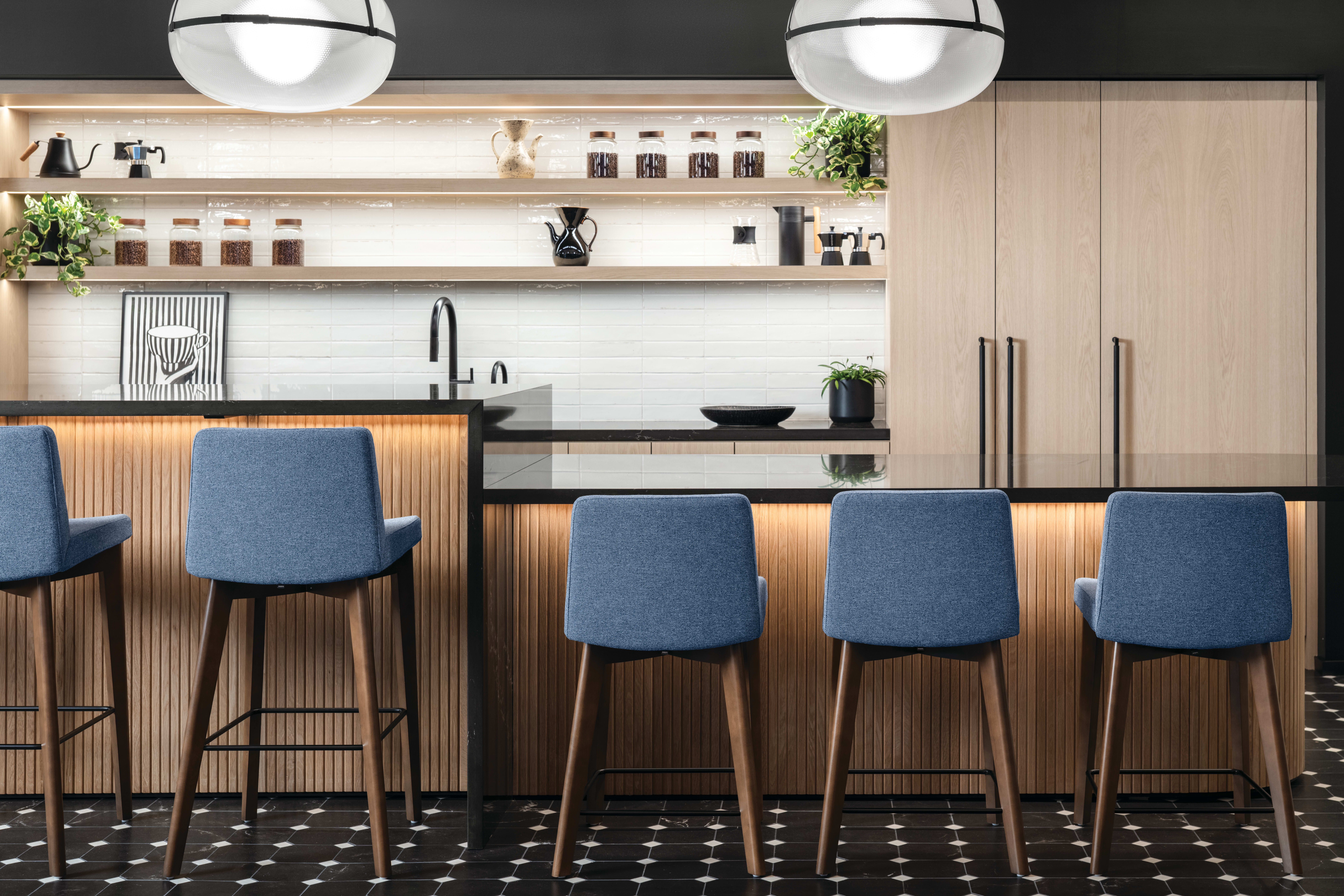 Cafe-style bar area with wood slat-front counter, black stone countertop, upholstered bar stools with wood legs, open shelving, and tall storage cabinets.