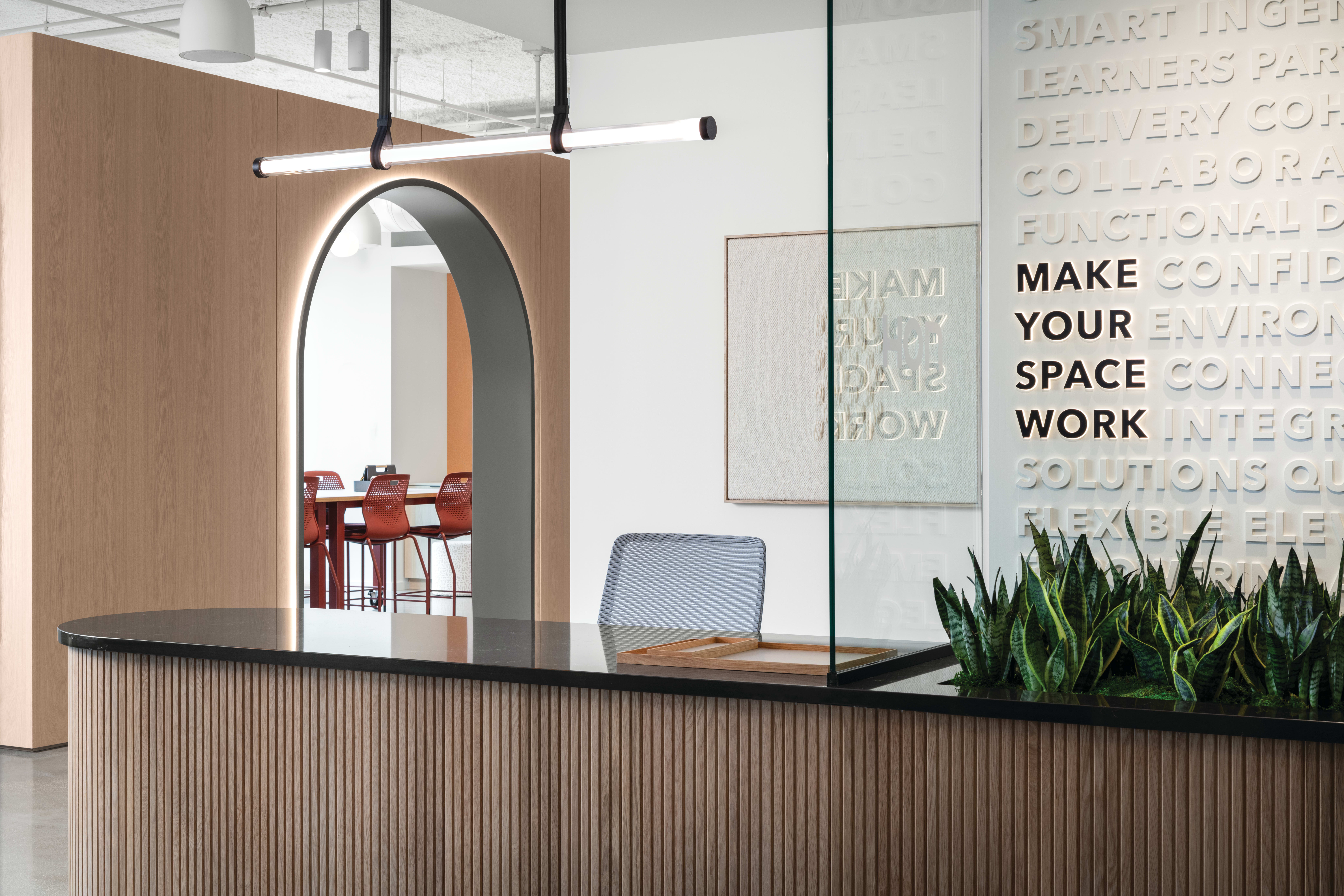 Reception counter with wood slat detailing and solid surface top, paired with task seating, integrated planter, pendant lighting, glass partition, and arched wall opening leading to collaborative seating area.